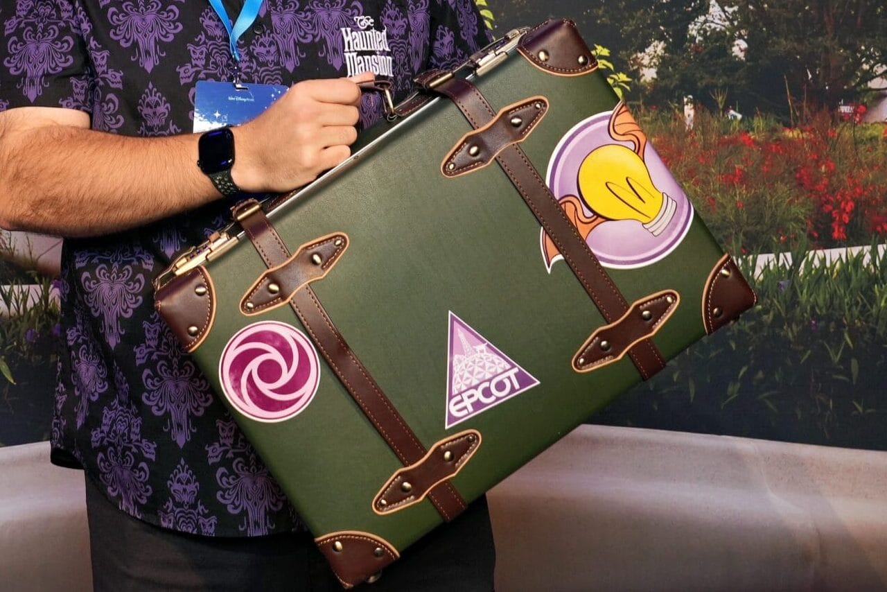 A man in a patterned shirt holds a decorated suitcase by Epcot's sphere during the EPCOT Food & Wine Festival.