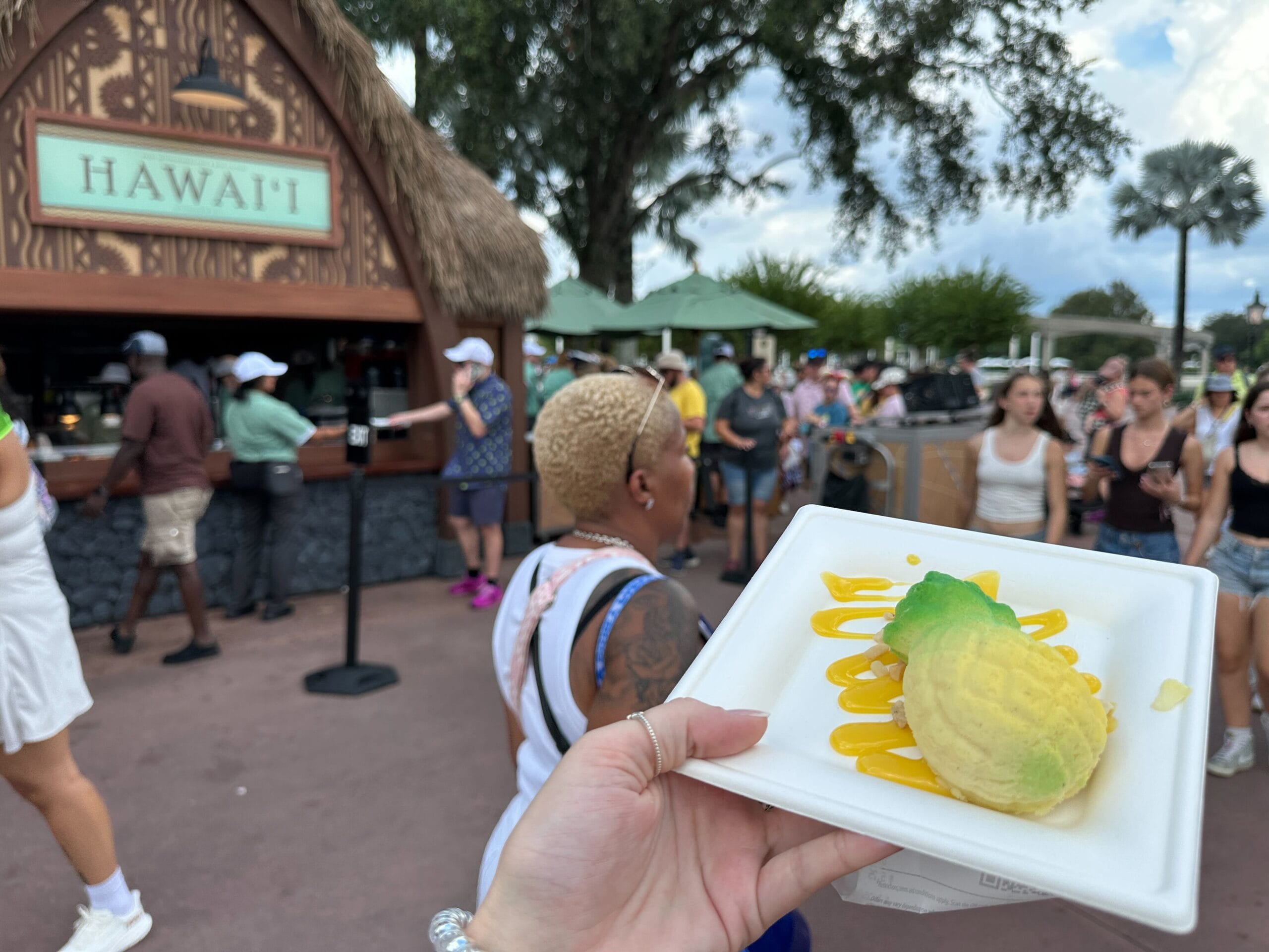 A hand holds a plate with a pineapple-shaped dessert at a bustling Food & Wine Hawaii booth during an outdoor event.