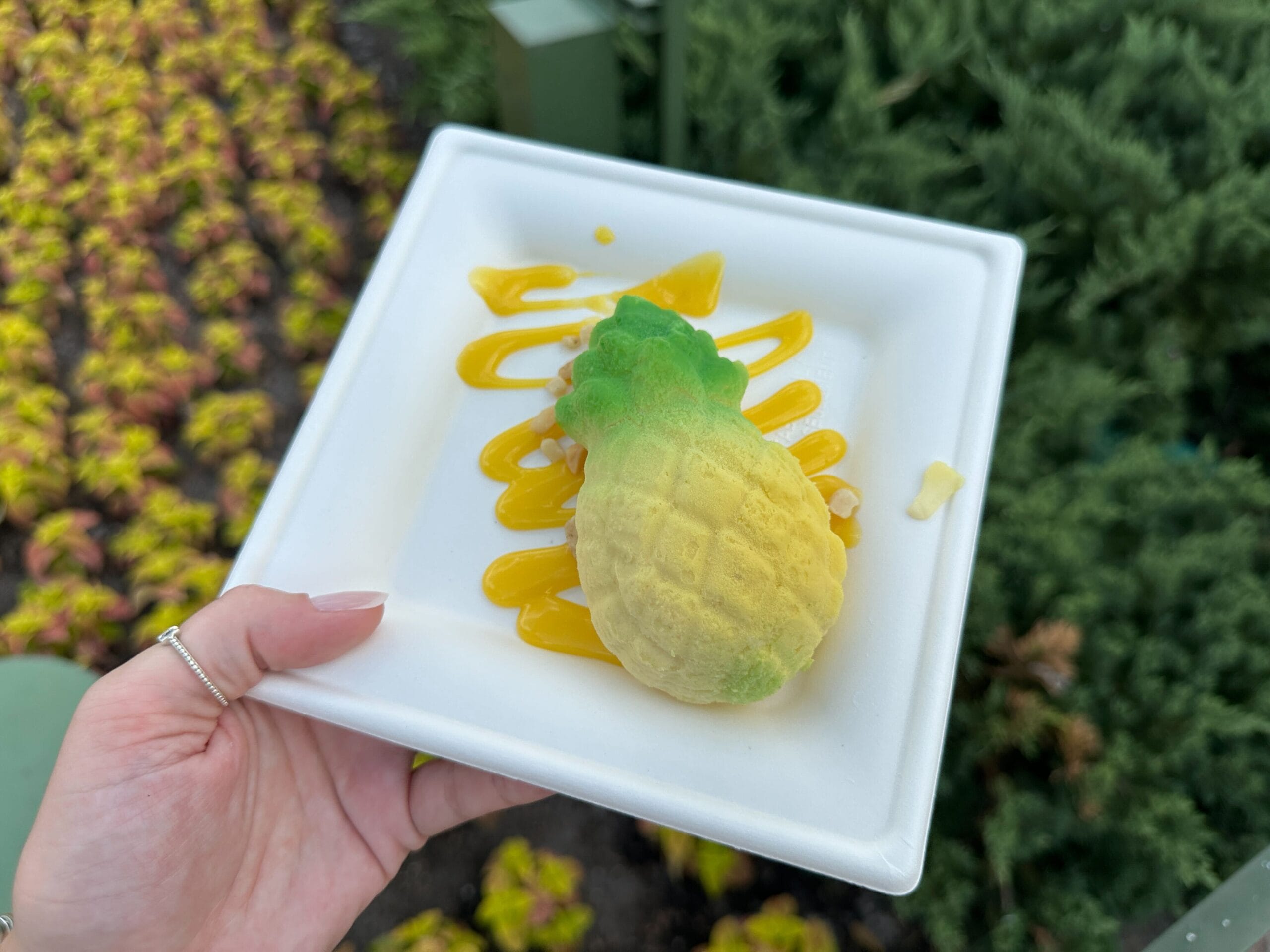 A hand holds a white plate with a pineapple-shaped dessert, perfect for Food & Wine lovers, greenery in the background.