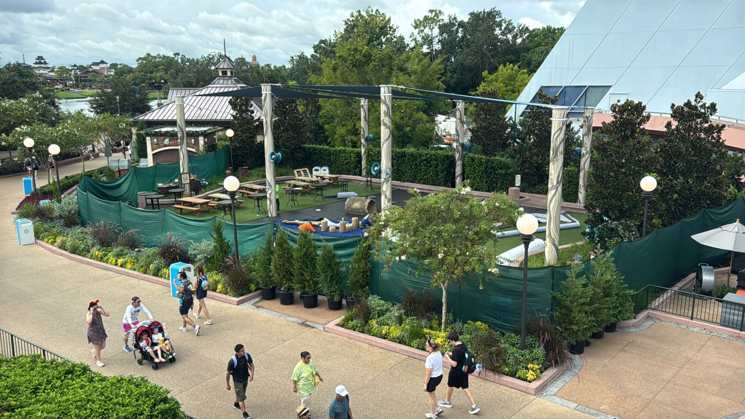 People walk past a fenced-off outdoor area under construction with plants, tables, and green barriers on a cloudy day.
