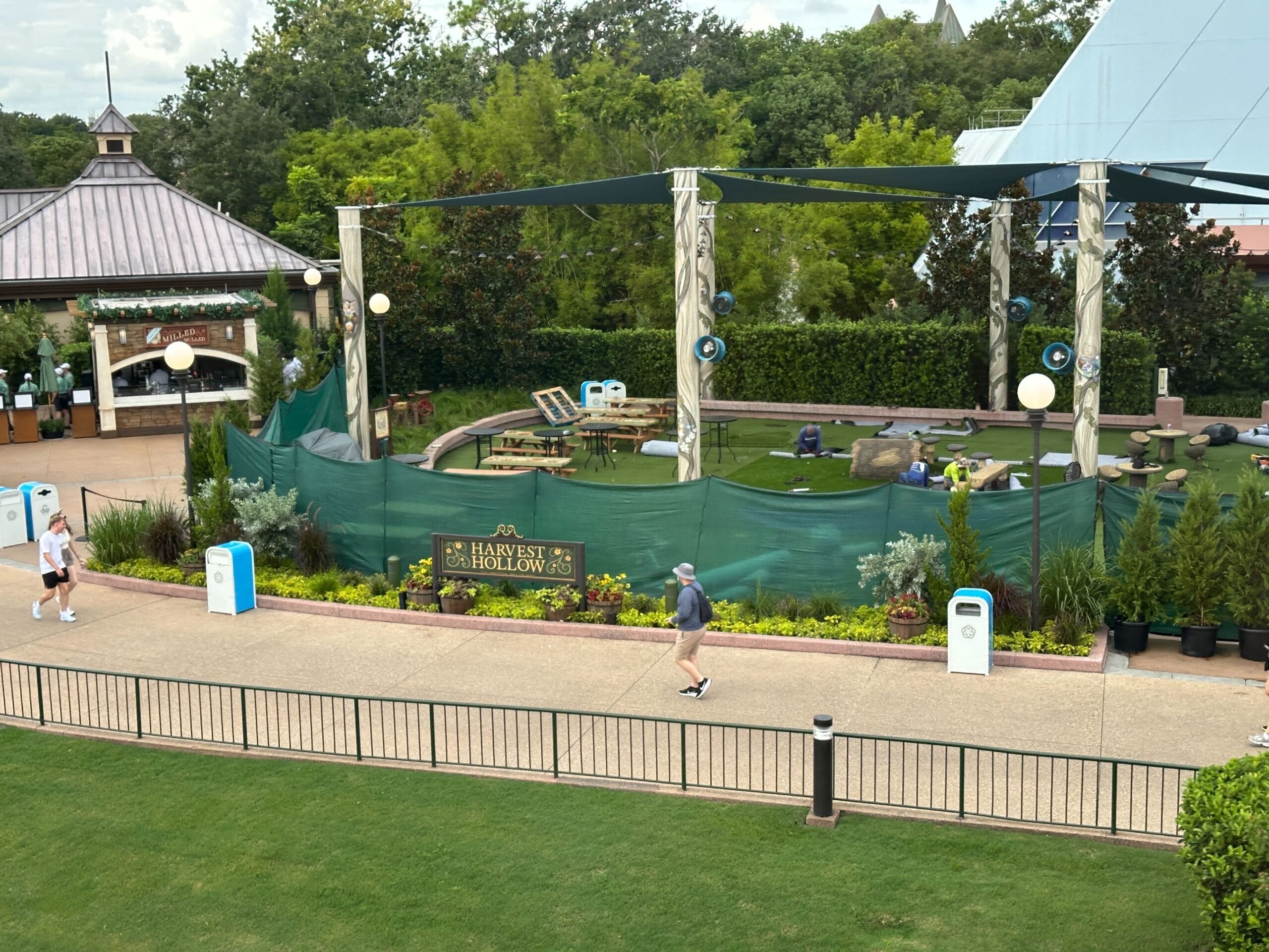 A fenced-off area labeled "Harvest Hollow" with plants, benches, and people walking nearby in a park setting.
