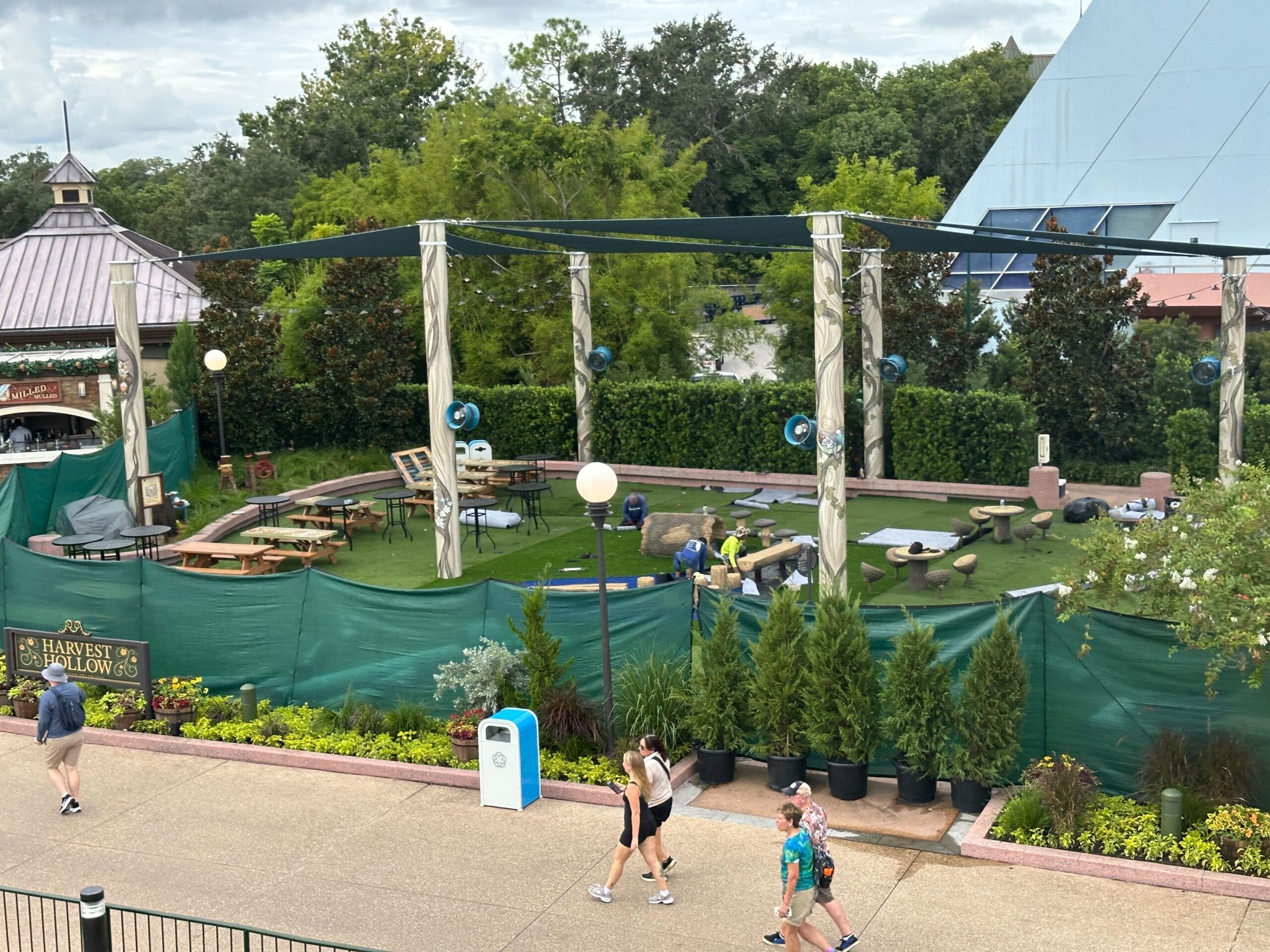 View of a construction area with green fencing, potted plants, and people walking by on a cloudy day.