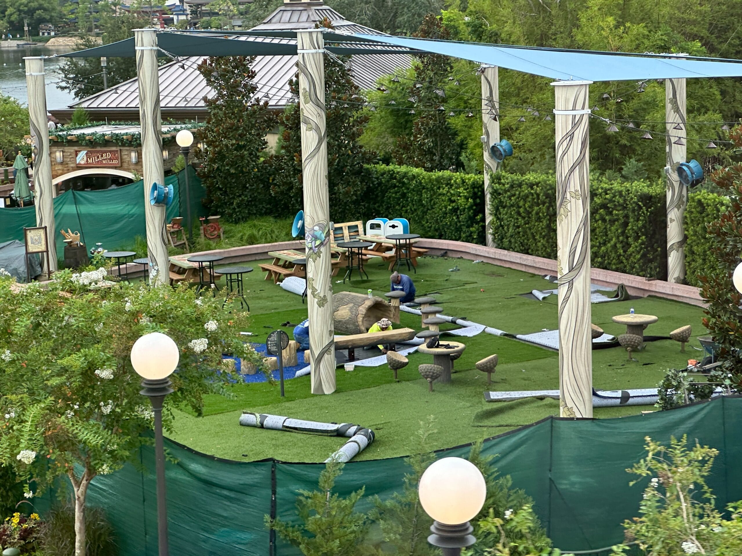 Mini golf course under construction with rolled turf, scattered equipment, and decorative pillars surrounded by greenery.
