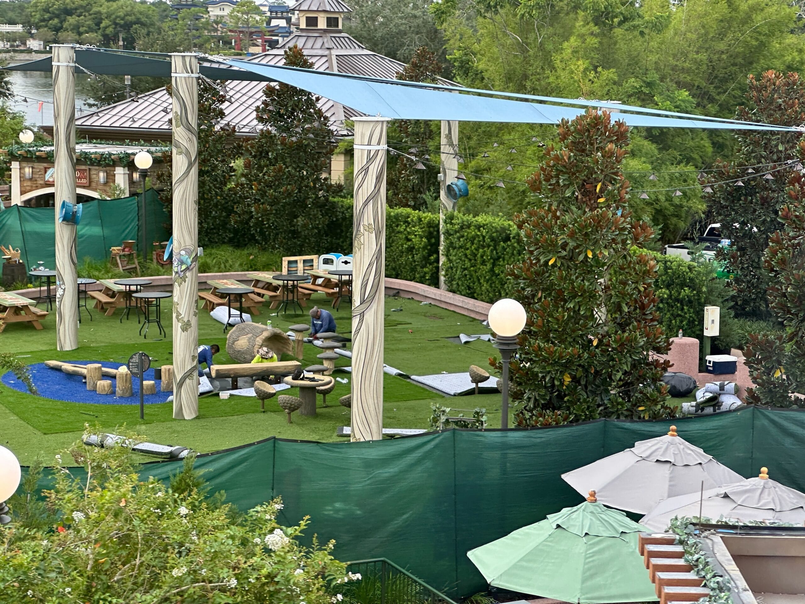 A fenced outdoor play area with artificial turf, benches, tables, and large umbrellas, seen from above.