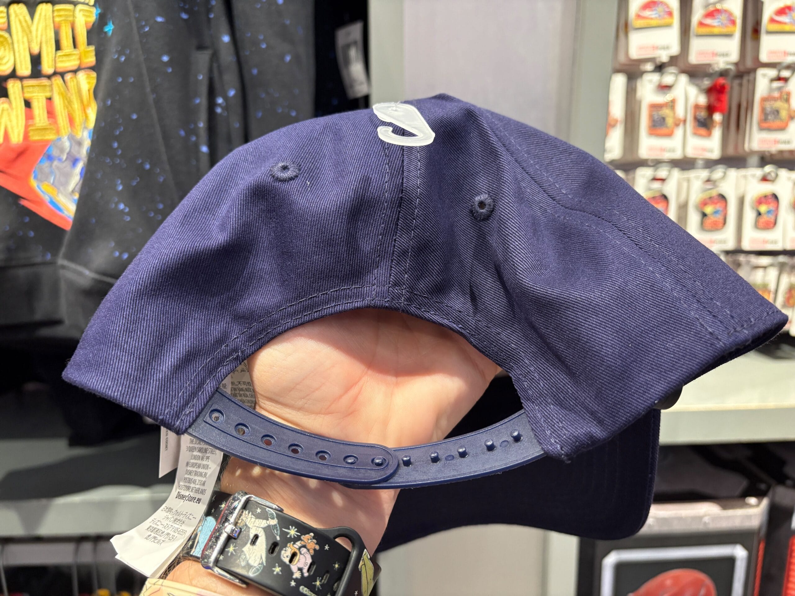 Hand holding a navy blue snapback hat, showing the adjustable strap and part of a store display in the background.