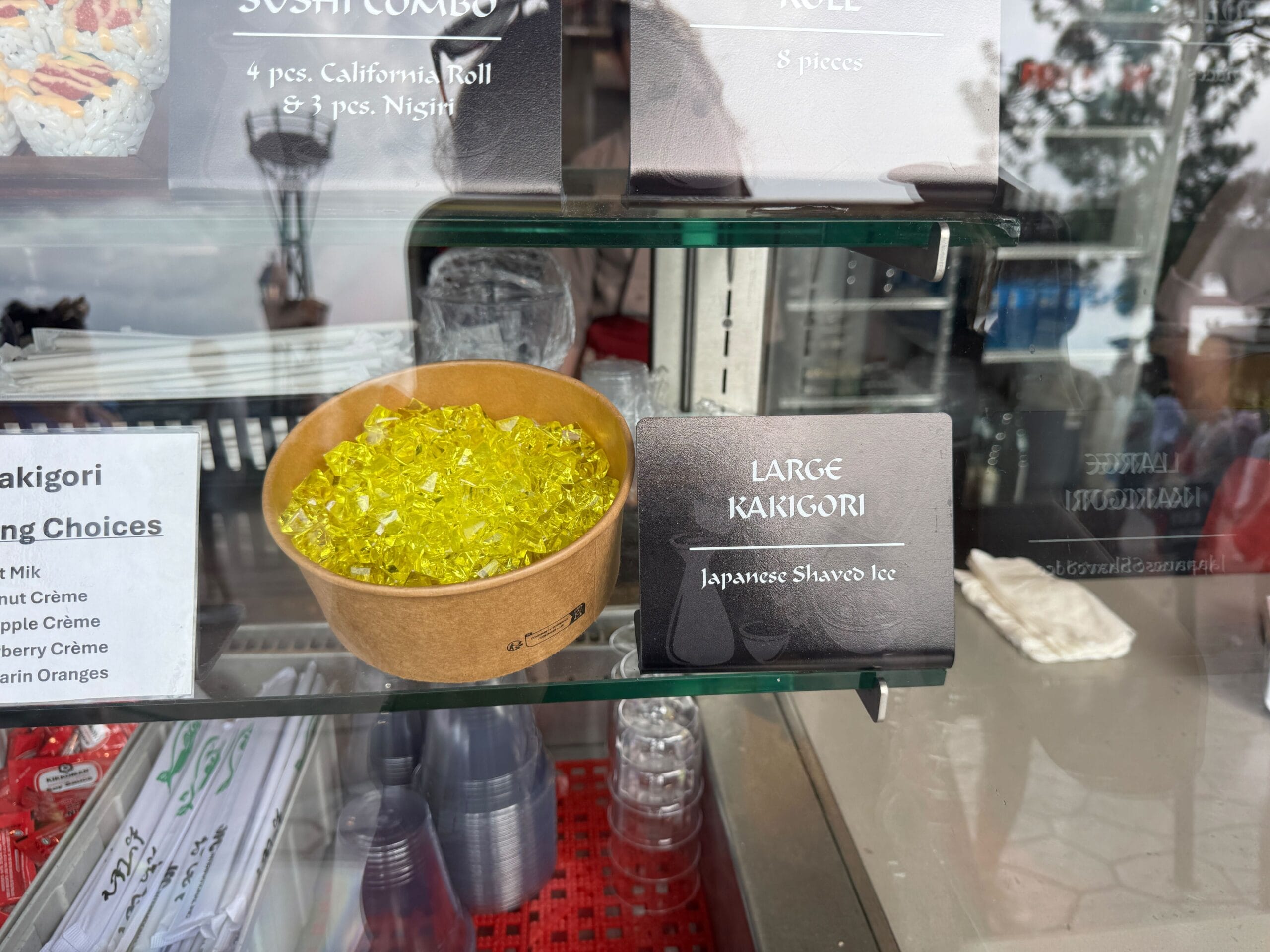 A bowl of yellow shaved ice sits beside a sign reading "Family-Sized Kakigori Japanese Shaved Ice" on a counter.