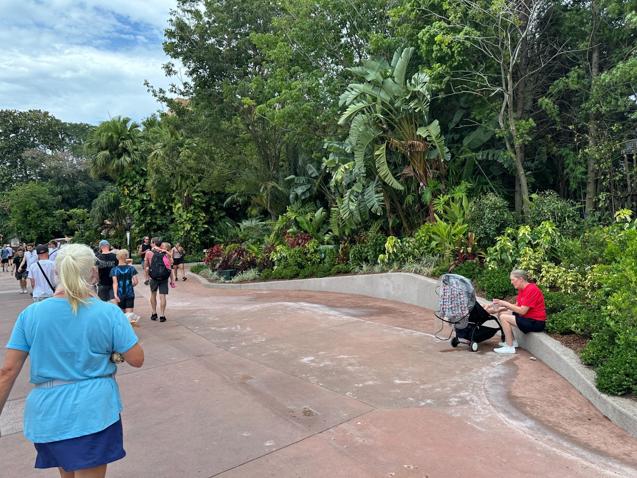 People walking on a paved path in a lush, green park; a man sits on a bench with a stroller nearby.
