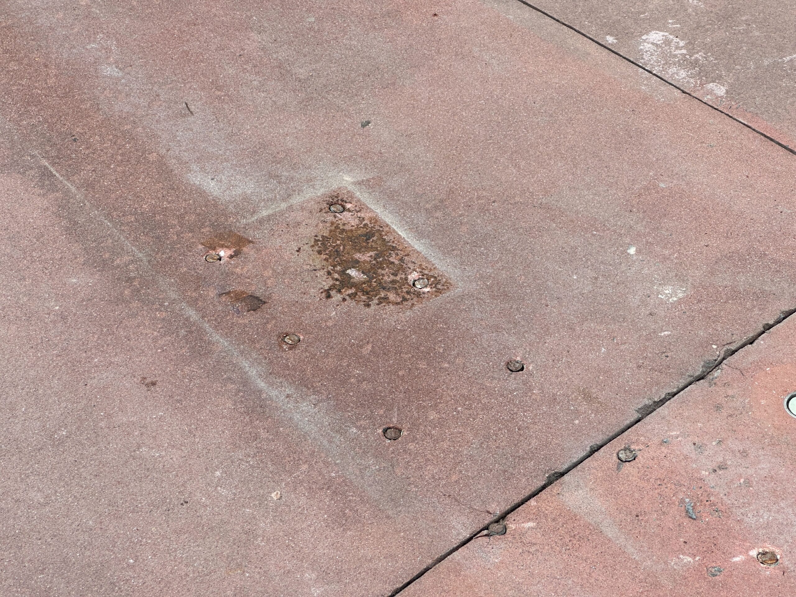 Reddish-brown concrete pavement with bolts and a rust-stained rectangular patch in the center.