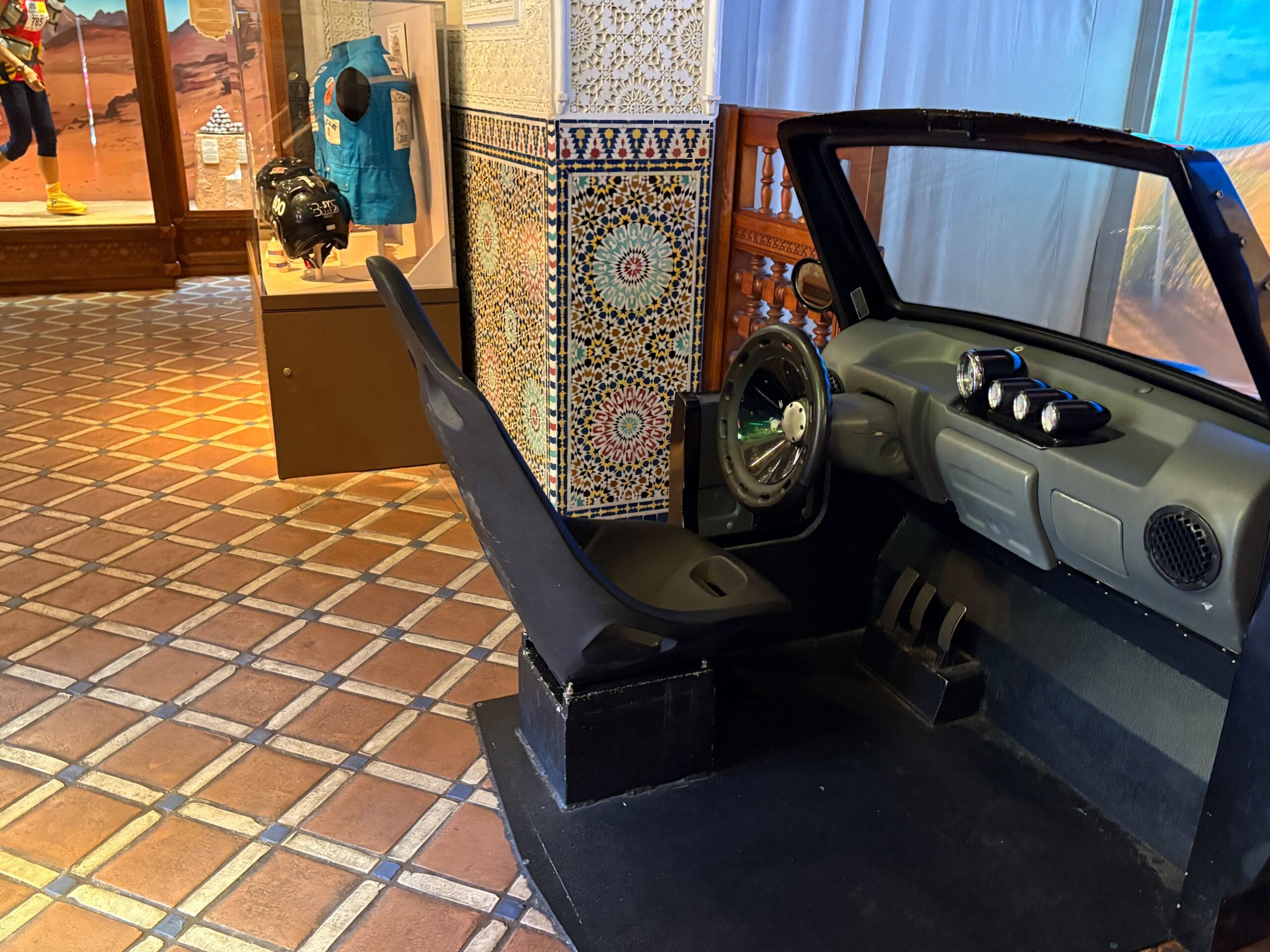 Car cockpit display with single seat and steering wheel in a tiled room with museum exhibits in the background.
