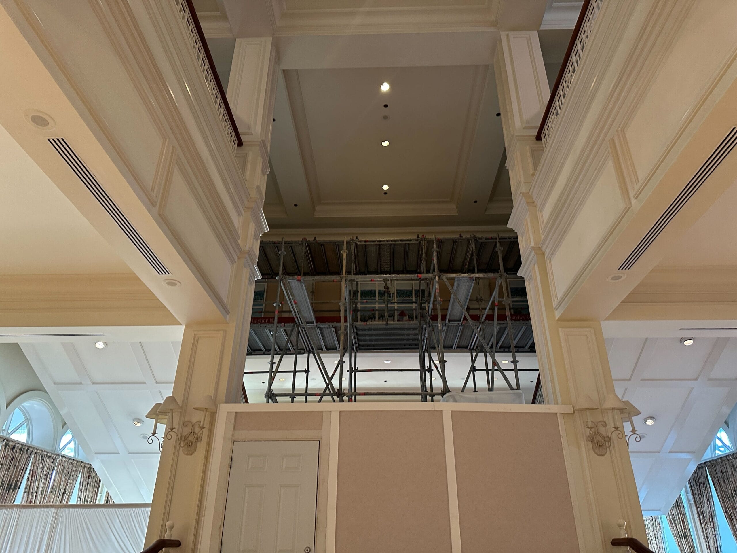 Scaffolding and construction barriers inside a large, high-ceilinged room with cream-colored walls and decorative lights.