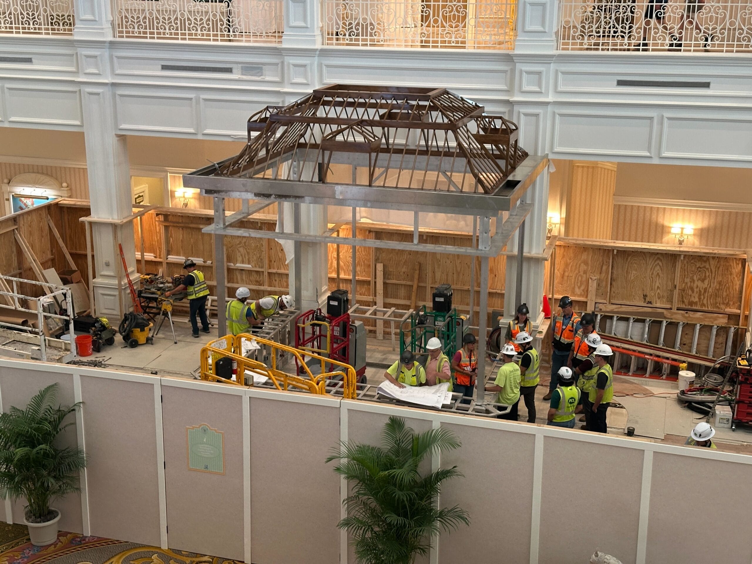 Construction workers in safety gear work under a partially built roof structure inside a large, elegant indoor space.