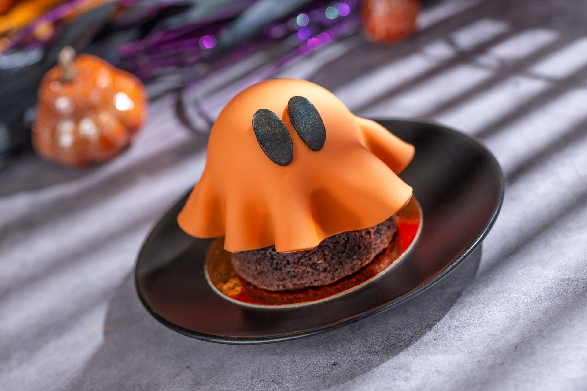 Chocolate dessert covered with orange fondant shaped like a cute ghost, on a black plate with Halloween decorations.