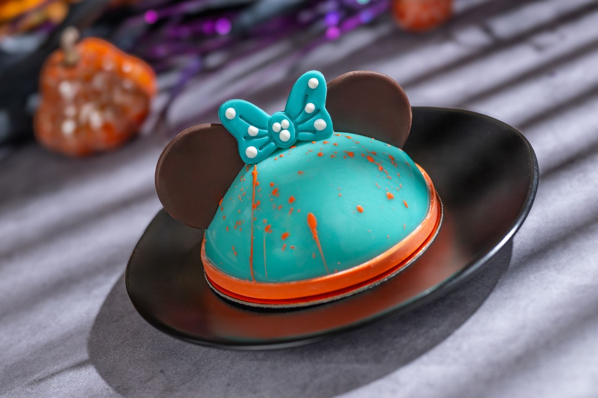 A dome-shaped teal dessert with chocolate ears and a bow, on a black plate, Halloween decorations in the background.