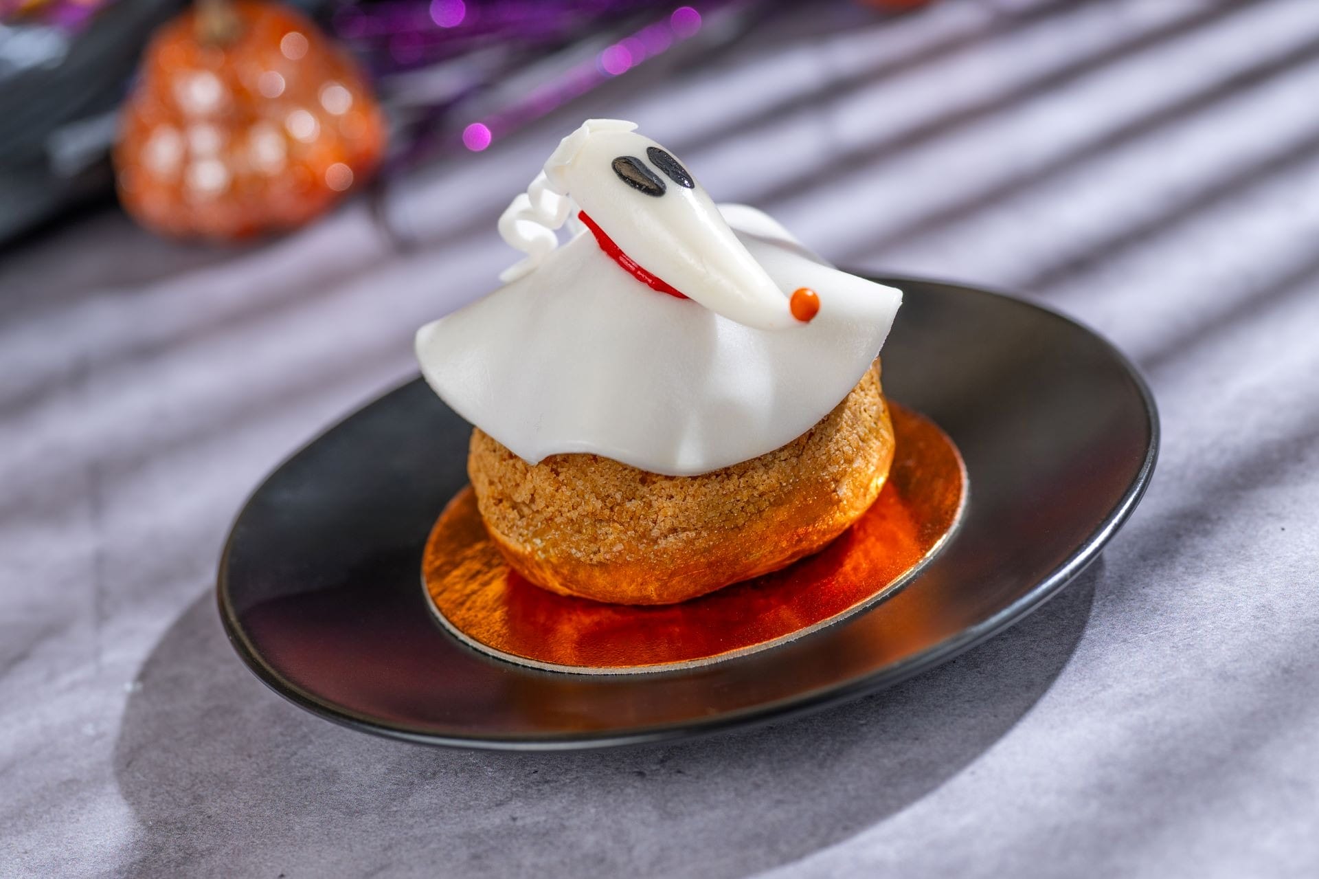 A pastry topped with white fondant shaped like a ghost, on a black plate with a gold base.