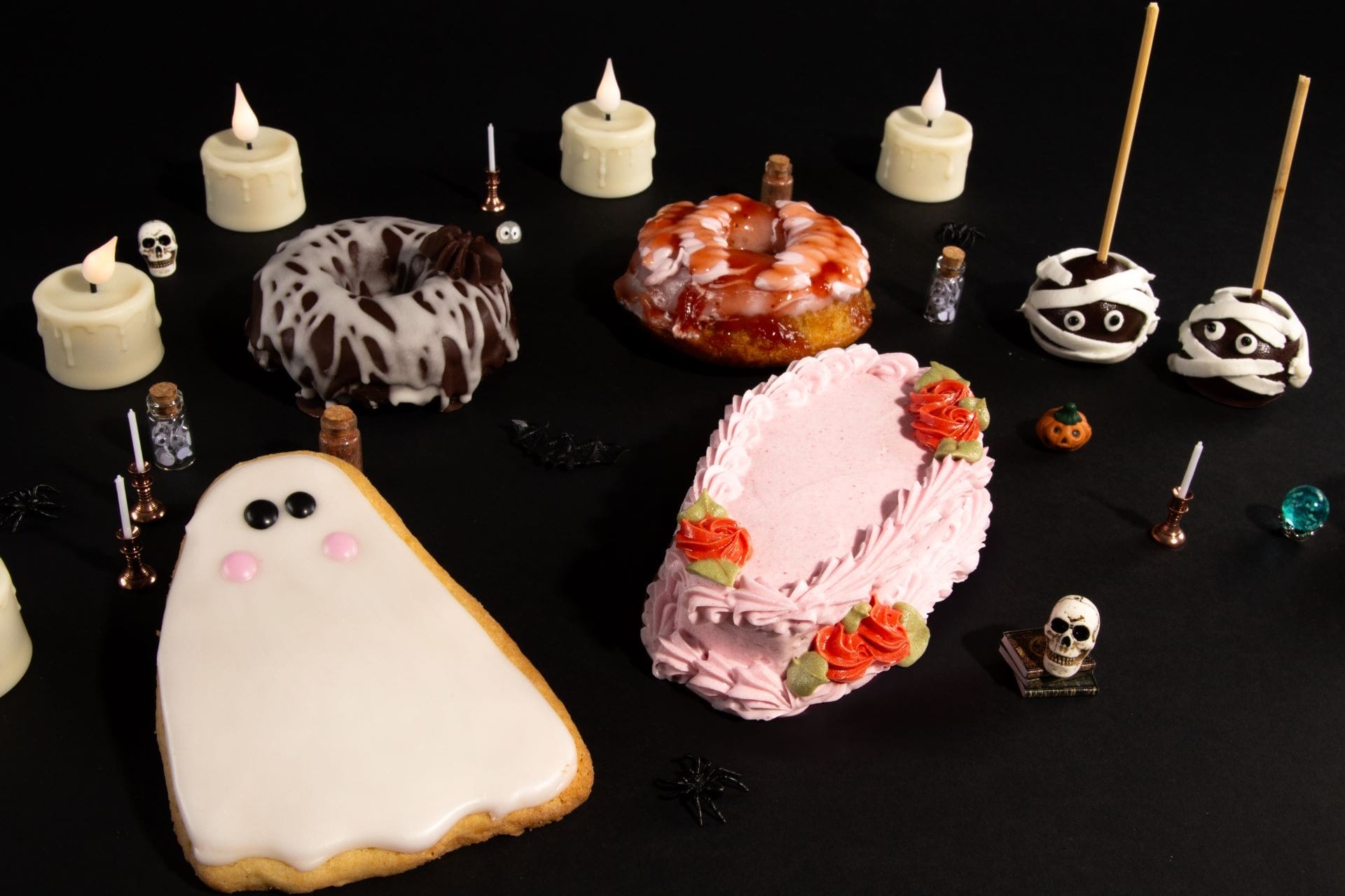 Halloween-themed desserts, including ghost and mummy cookies, cakes, candles, and small skeleton decorations on a black background.