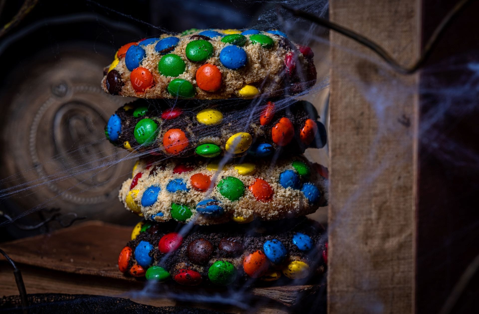 A stack of three cookies with colorful candy coating, surrounded by cobwebs, sits on a wooden surface.