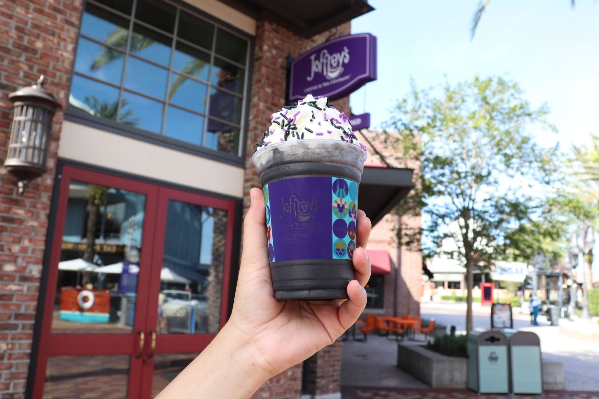 A hand holds a whipped cream-topped drink in front of Joffrey’s at Walt Disney World on a sunny day.