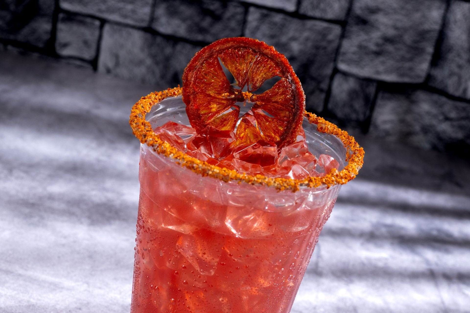 A red iced drink in a chili-rimmed glass, perfect for 2025 Halloween treats, topped with dried citrus by a stone wall.