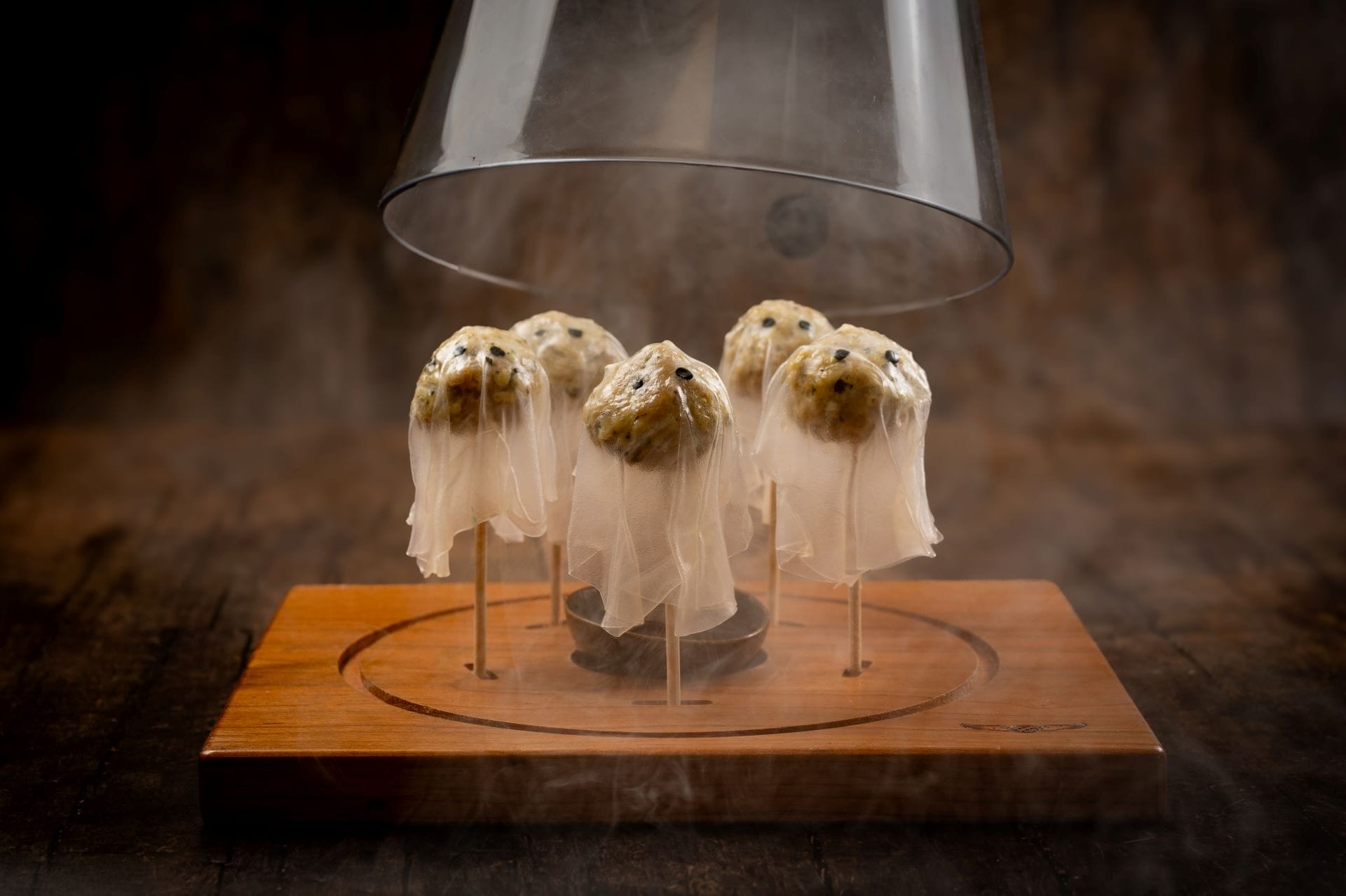 Five ghost-shaped treats on sticks, covered with thin white cloth, displayed under a glass dome on a wooden base.