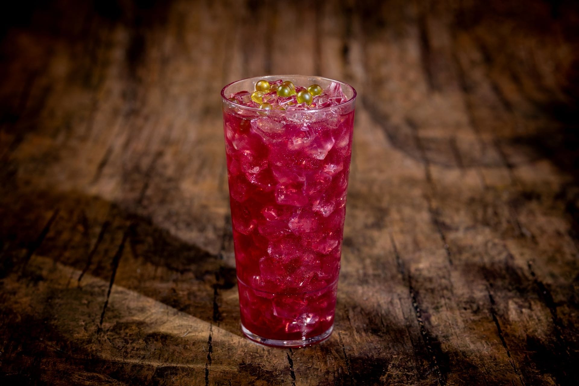 A tall glass of pink-red iced drink with small green berries on top, placed on a rustic wooden surface.