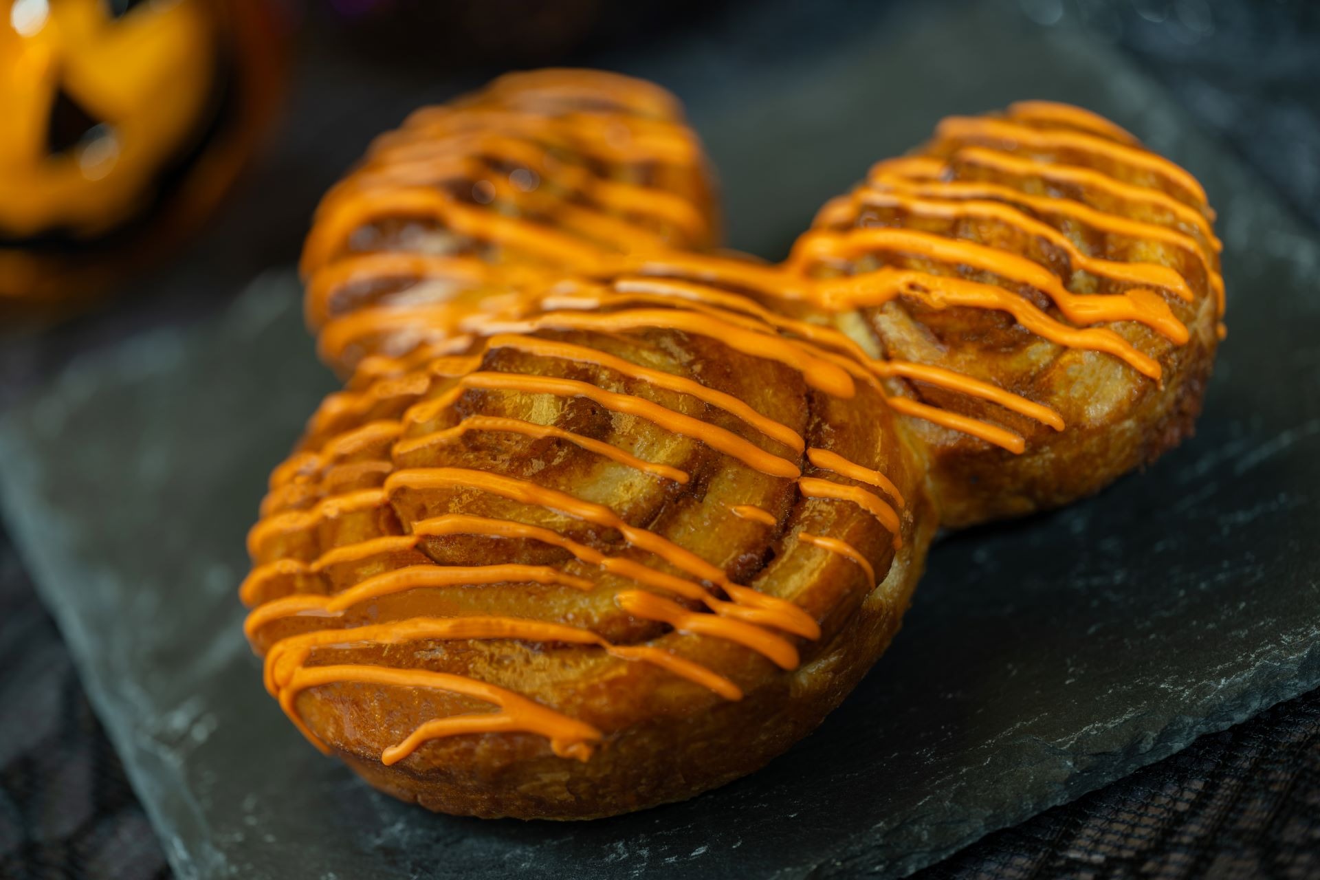 Three cinnamon rolls with orange icing drizzle arranged on a dark slate surface—perfect for 2025 Halloween Treats.