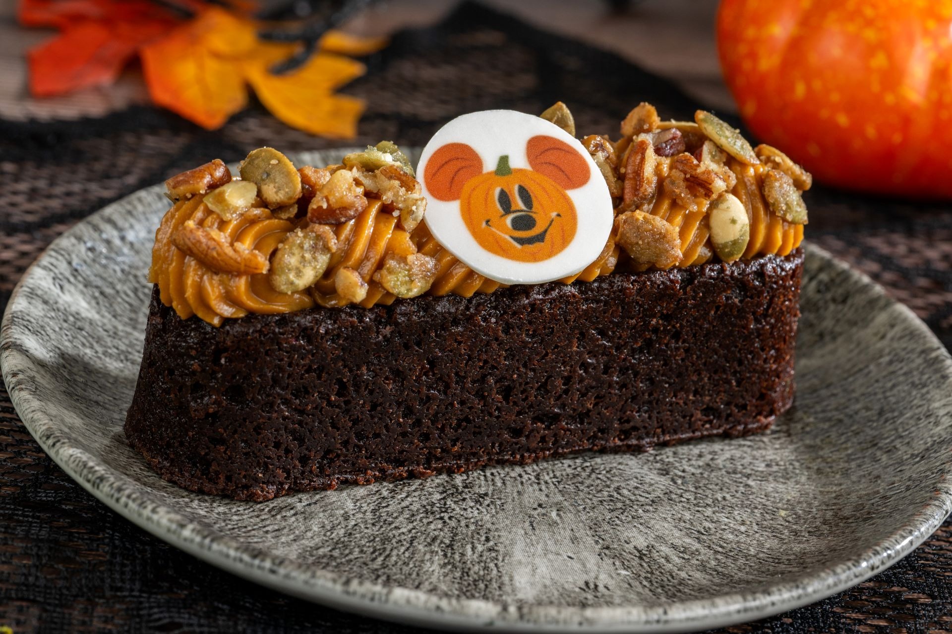 A slice of chocolate cake with orange frosting, nuts, and a pumpkin Mickey atop a gray plate—part of 2025 Halloween Treats.