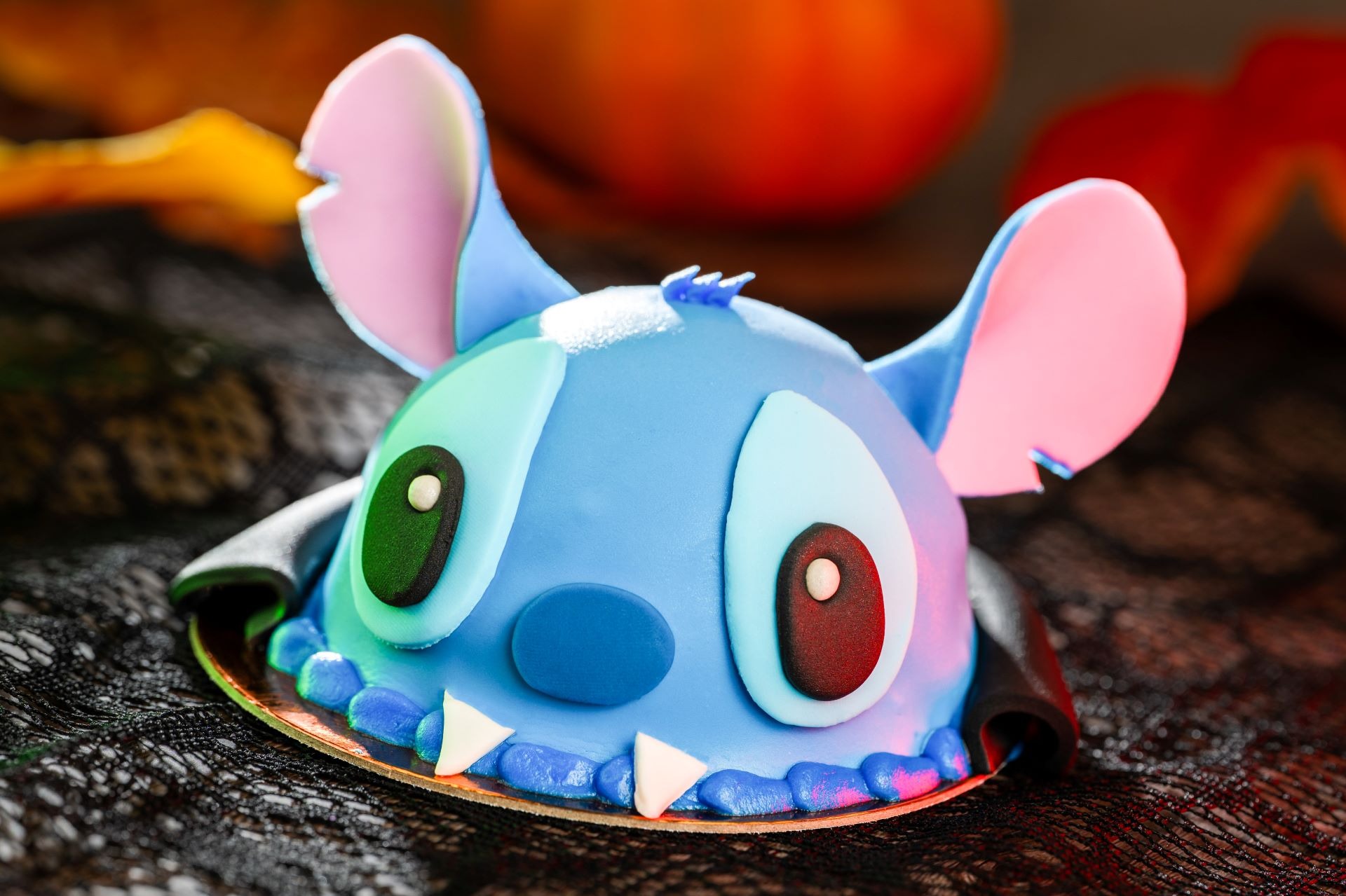 A round cake decorated to look like Stitch from Walt Disney World, with large blue ears and colorful eyes.