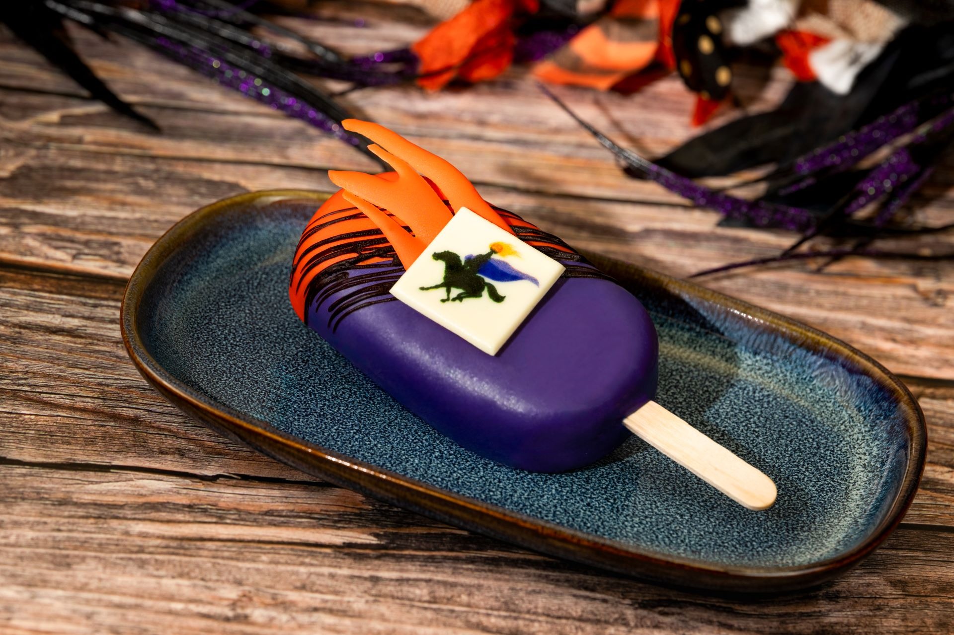 A purple popsicle with orange and black decorations, inspired by 2025 Halloween Treats, on a blue plate.