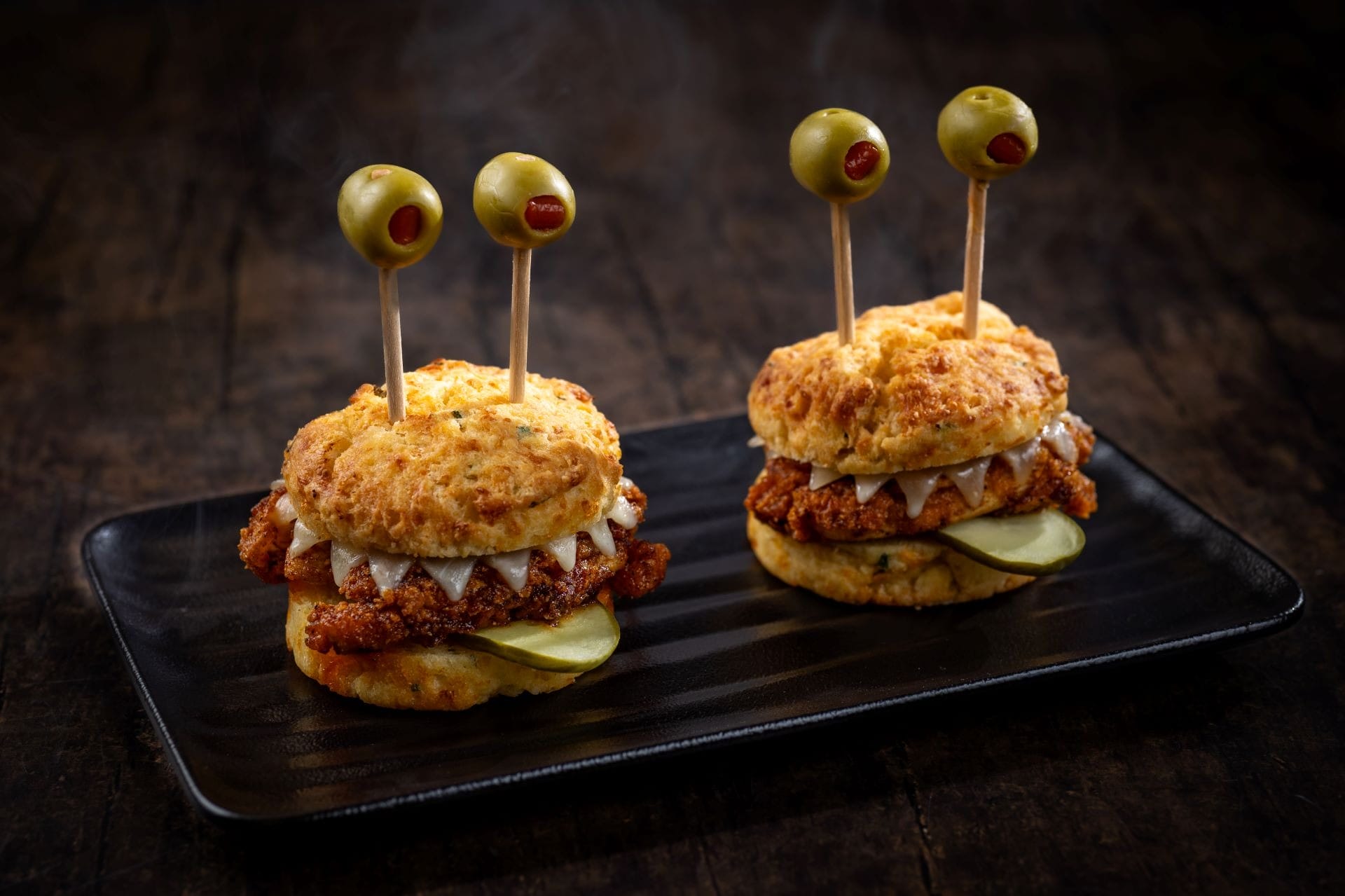 Two sandwiches decorated as monsters make fun 2025 Halloween Treats, served with olive eyes on a black plate.