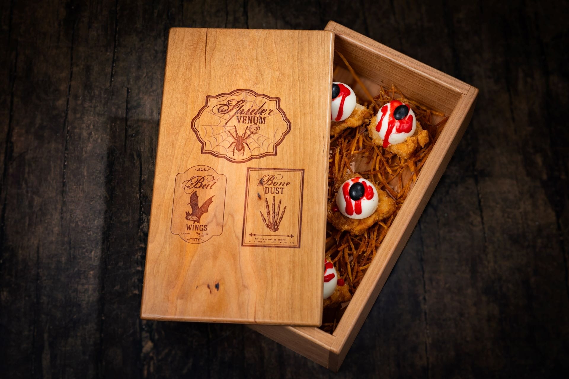 A wooden box partly open, revealing 2025 Halloween treats with spooky white frosting and red and black decorations inside.