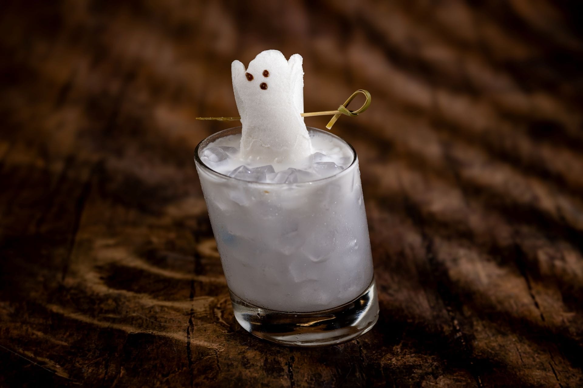 A cocktail with ice and a marshmallow ghost garnish on a wooden surface.
