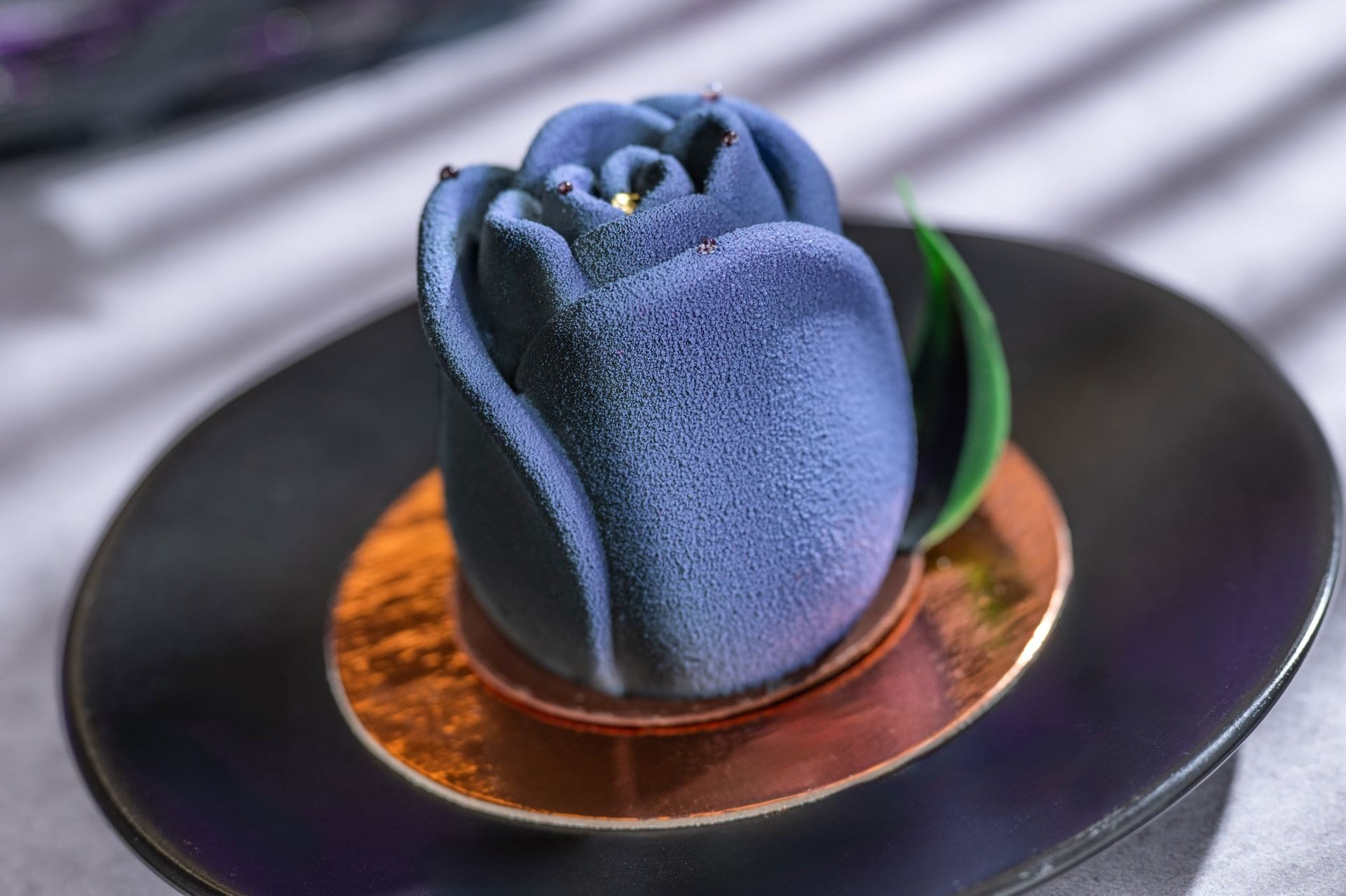A blue, rose-shaped mousse dessert with a green leaf on a black plate and a gold base.