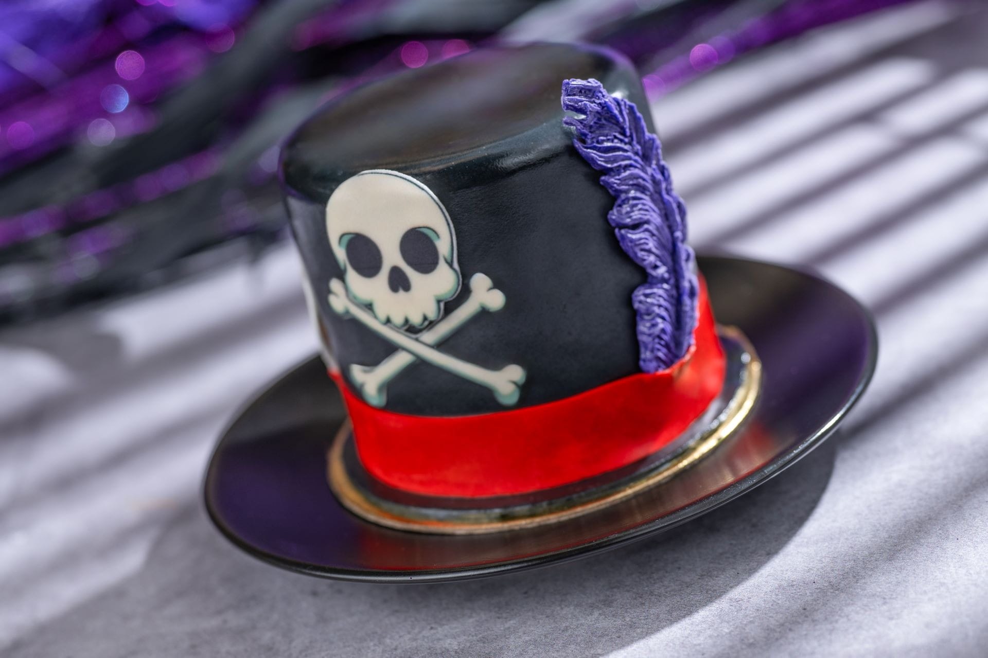 Black top hat cake with a skull and crossbones, red ribbon, and purple feather decoration on a black plate.
