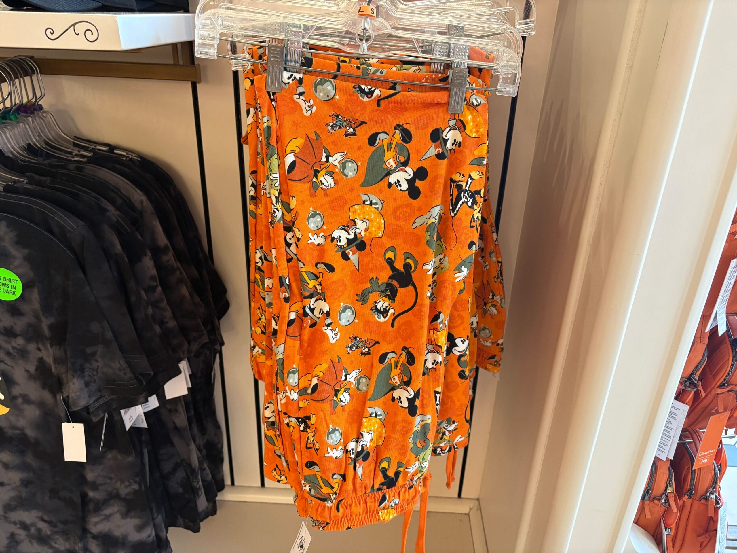 Bright orange pants with cartoon dog and pumpkin prints hang on a rack of Disney Halloween Merch in a clothing store.