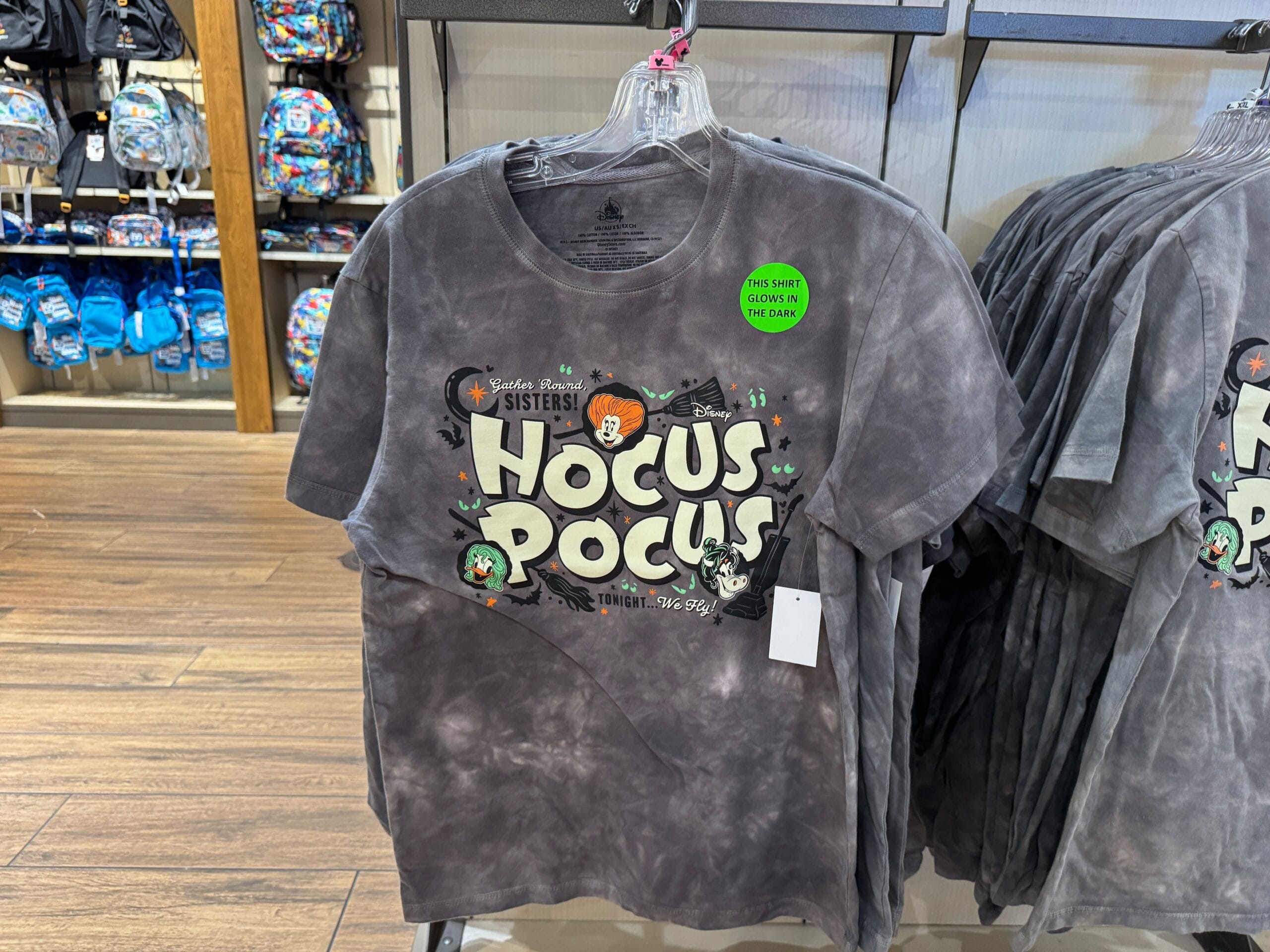 Gray "Hocus Pocus" t-shirt on a rack, part of Halloween merchandise, with a green "glows in the dark" sticker.