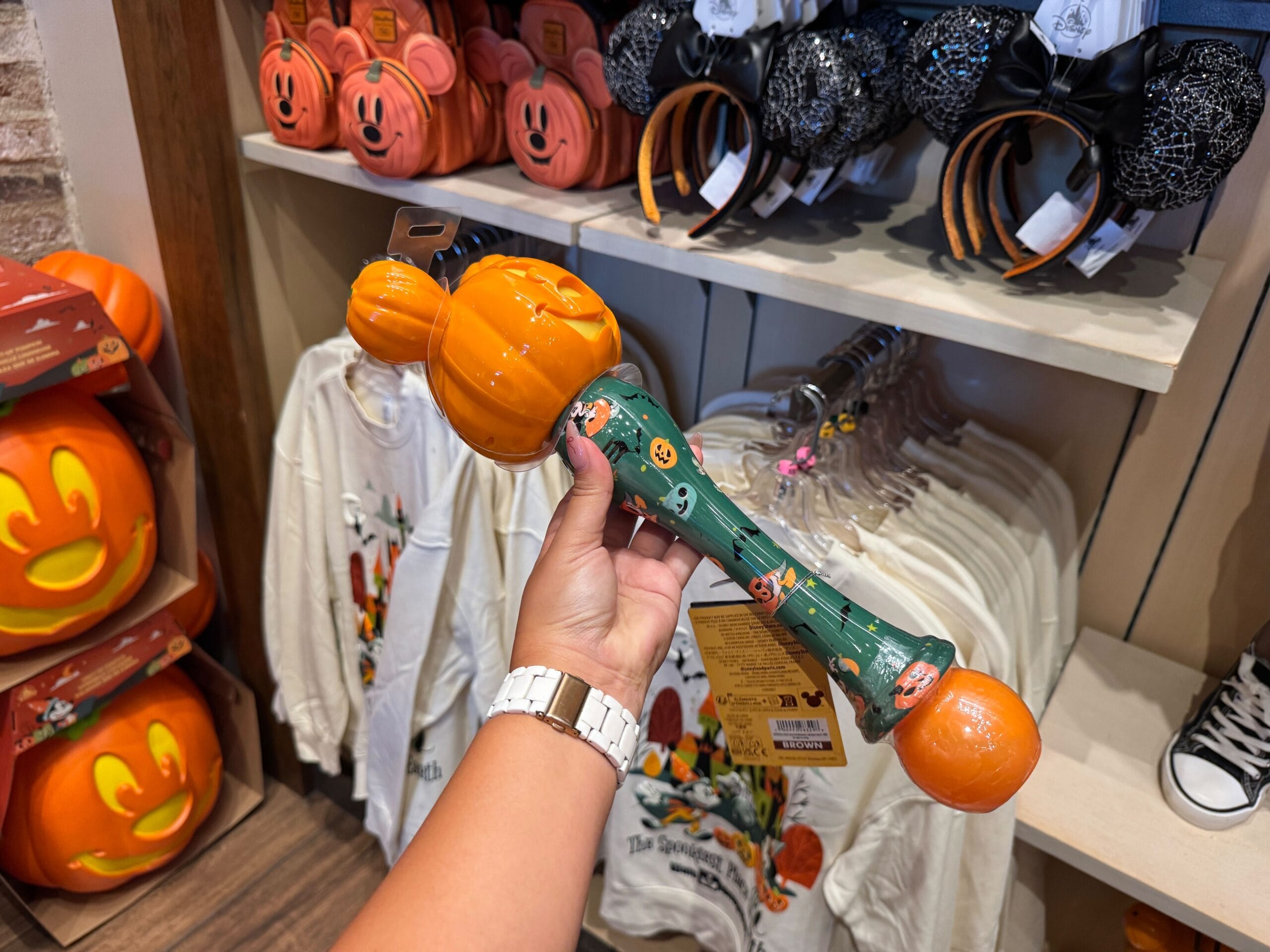 A hand holds a pumpkin-shaped wand in a store with Walt Disney World 2025 Halloween merchandise.