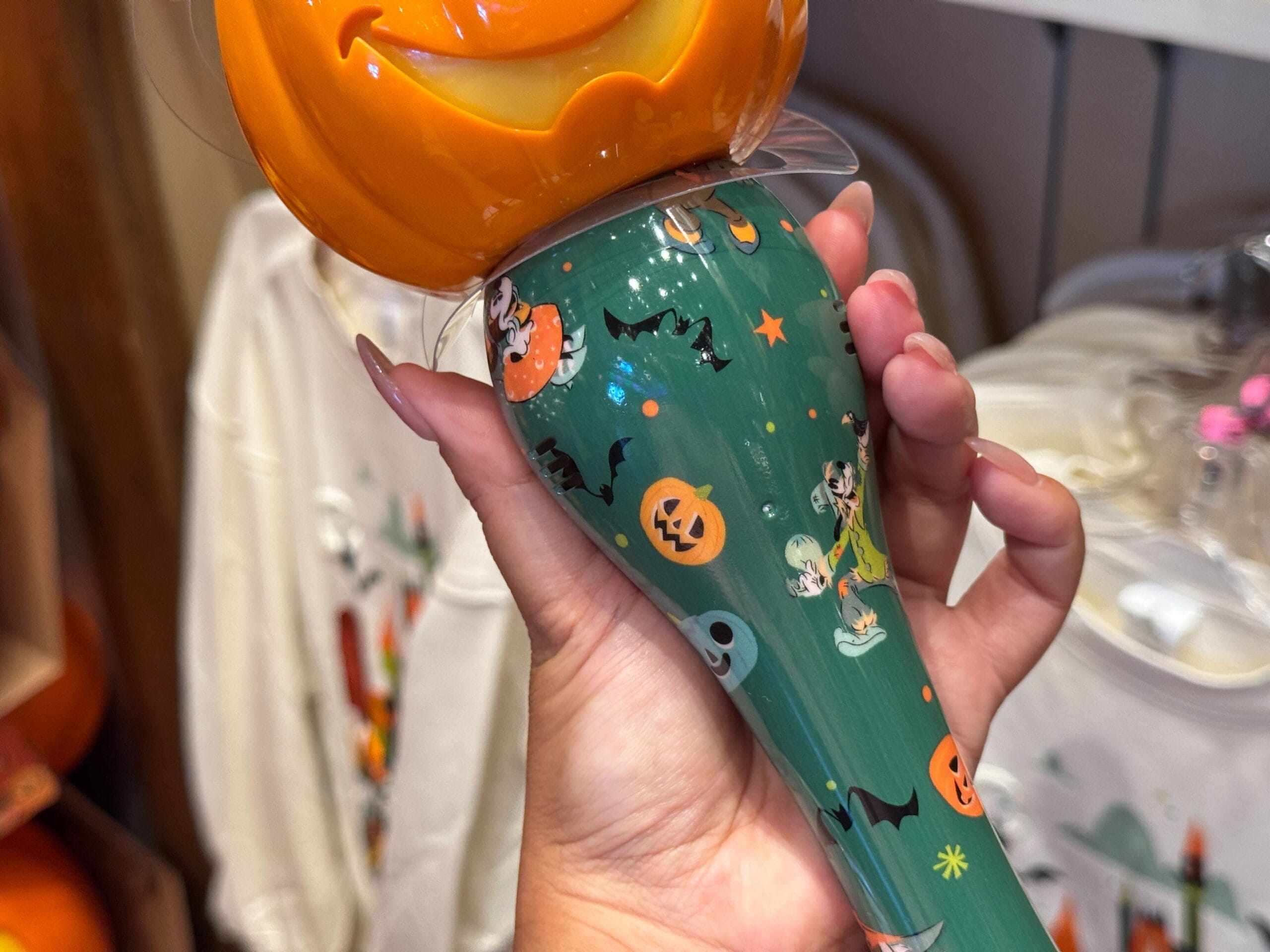 Hand holding a green 2025 Halloween Merchandise sipper cup with a smiling pumpkin lid and cartoon characters.