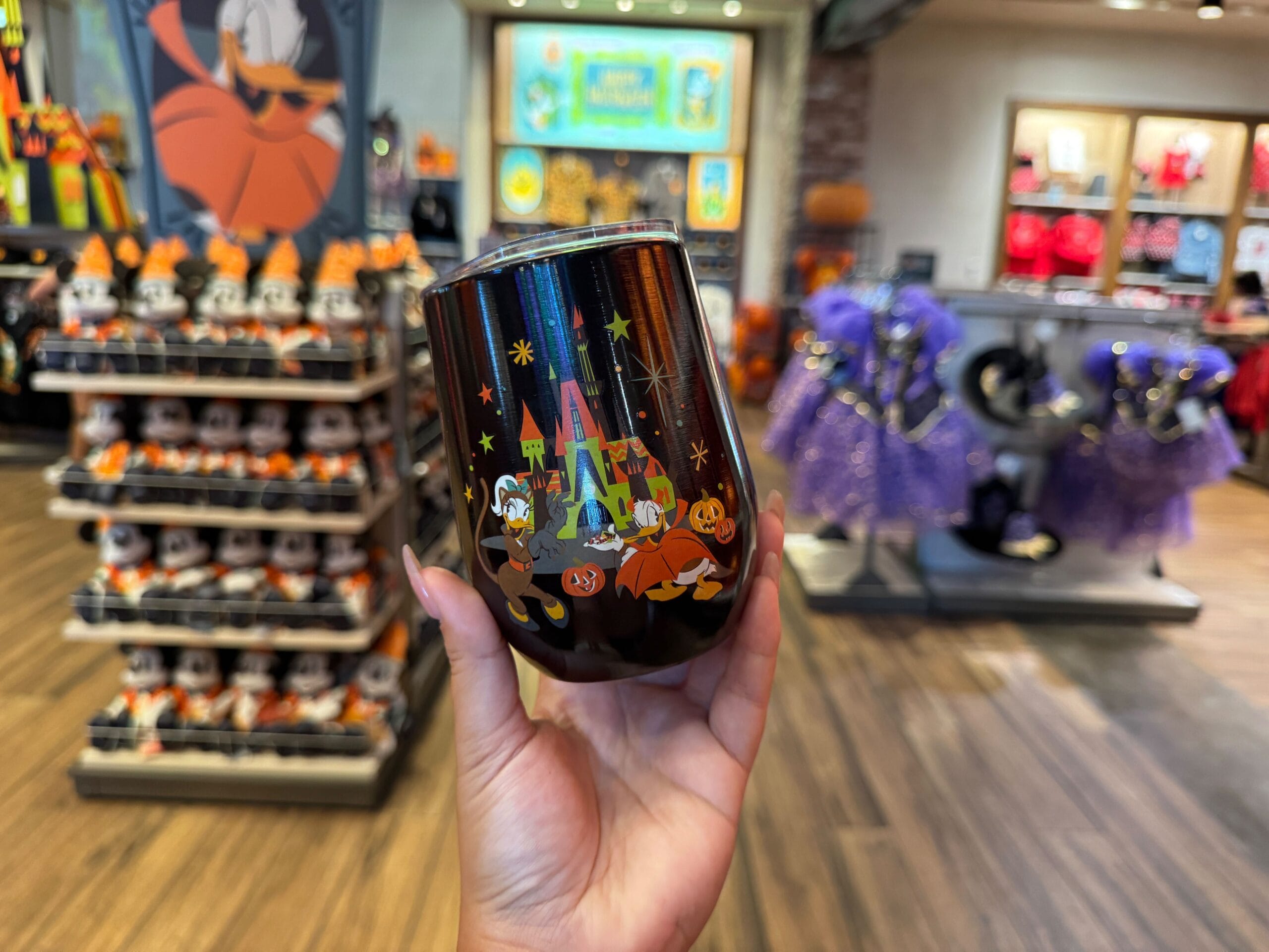Hand holding a Halloween-themed Disney tumbler amid Walt Disney World Halloween merchandise and costumes in the background.