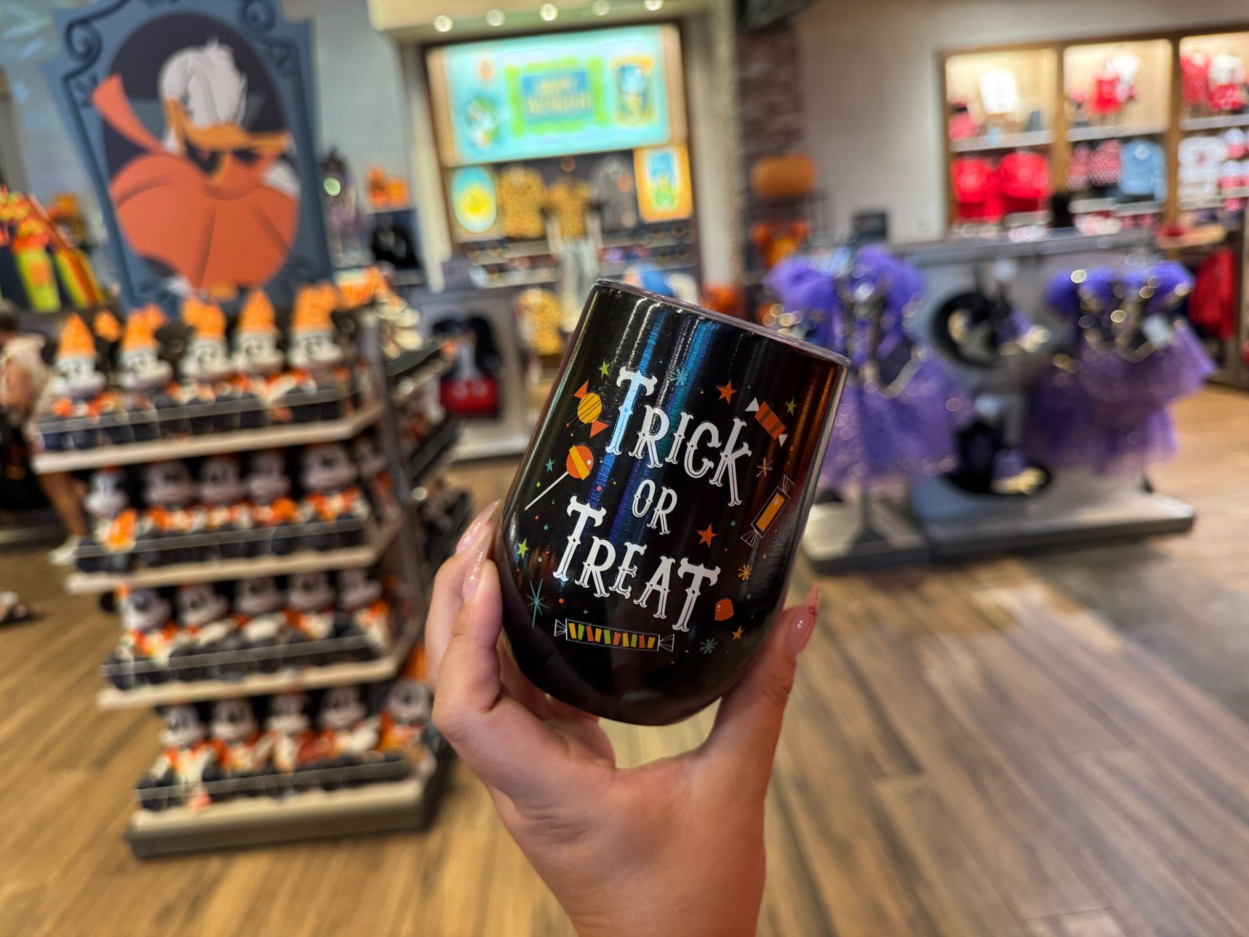A hand holds a black "Trick or Treat" cup among Halloween merchandise and costumes in a festive store at Walt Disney World.