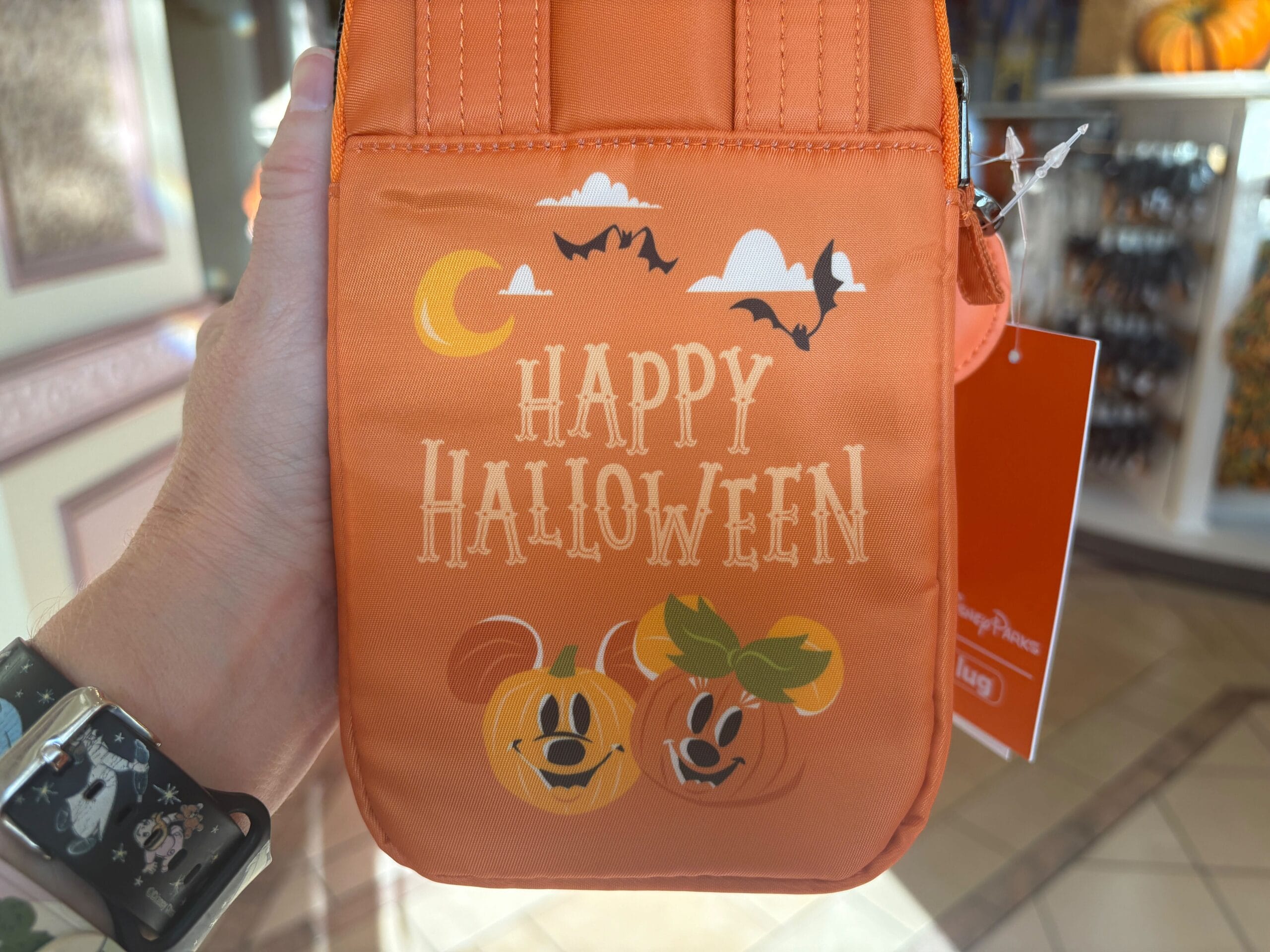 Orange bag with "Happy Halloween," bats, moon, pumpkins, held by a hand wearing a Walt Disney World-themed watch.