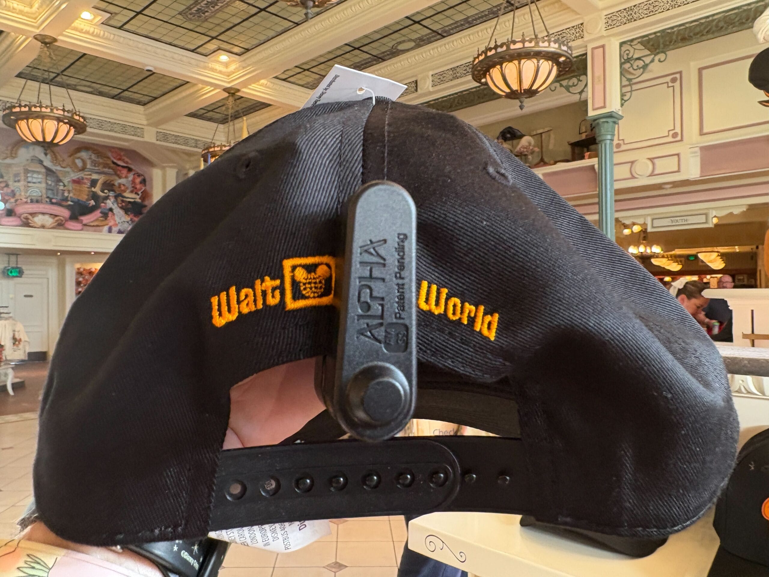 A hand holds a black "Walt World" cap with a security tag, surrounded by Halloween merchandise in the store.
