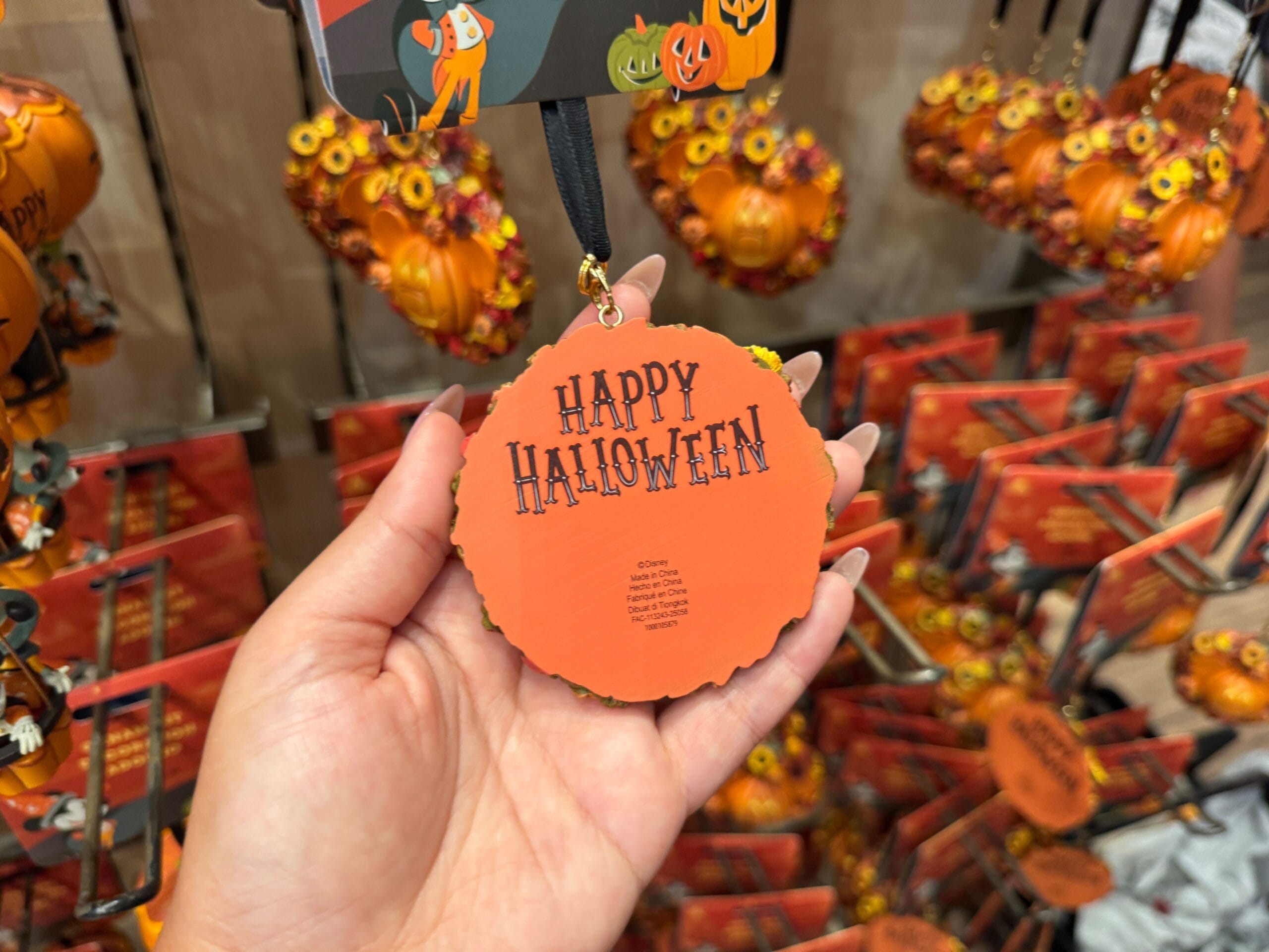 A hand holds a round "Happy Halloween" ornament, part of 2025 Halloween merchandise, in a store with festive decor behind.