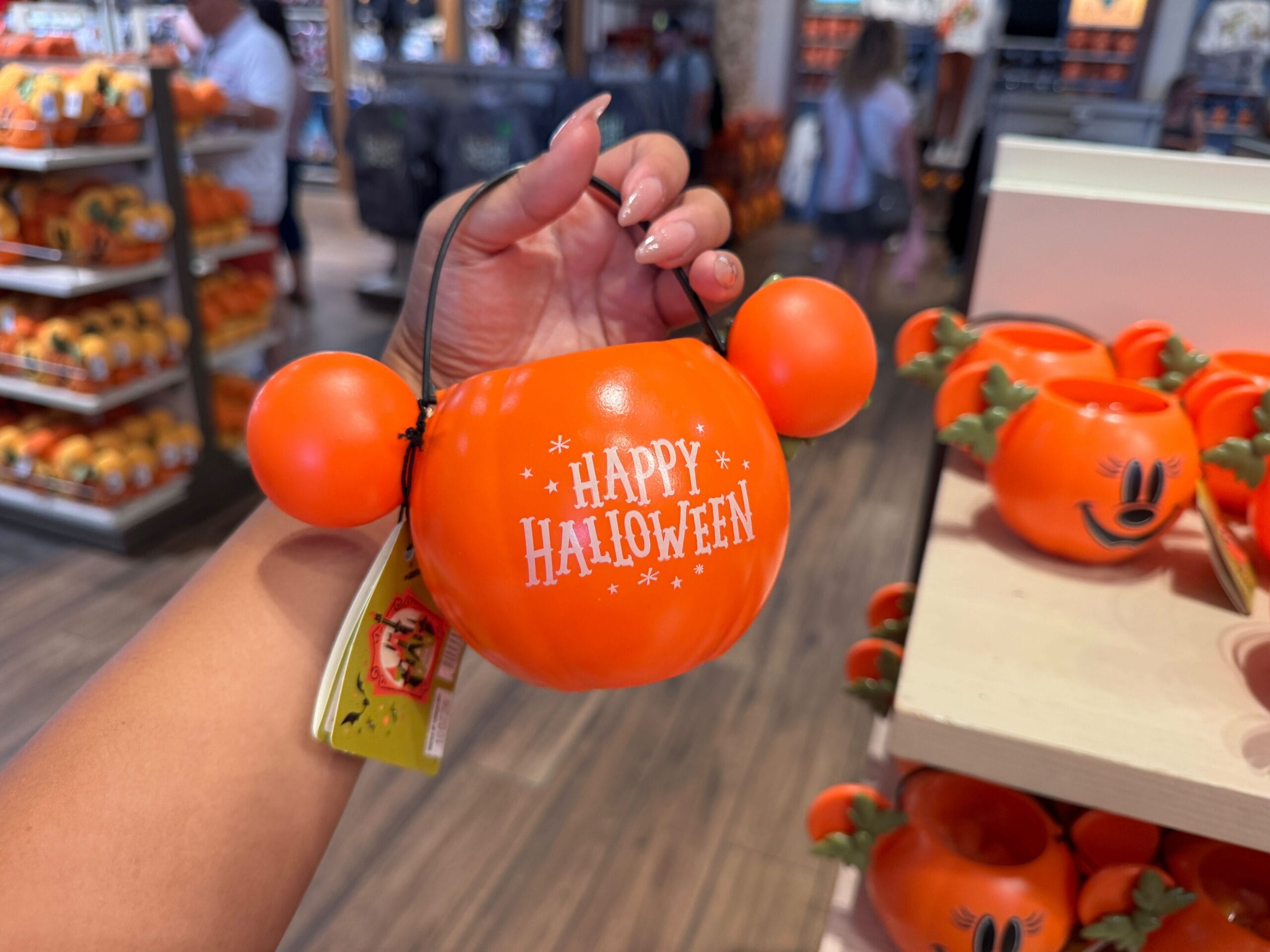 Hand holding a Mickey Mouse-shaped pumpkin that says "Happy Halloween" among 2025 Halloween Merchandise at Walt Disney World.
