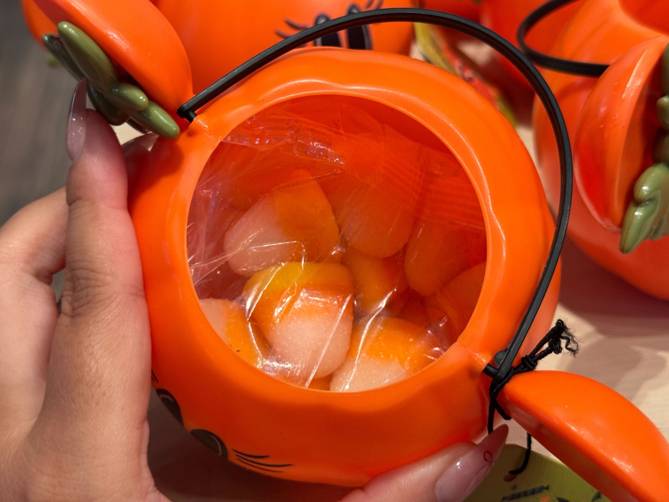 A hand holds a 2025 Halloween Merchandise pumpkin container with candy corn at Walt Disney World, wrapped in clear plastic.
