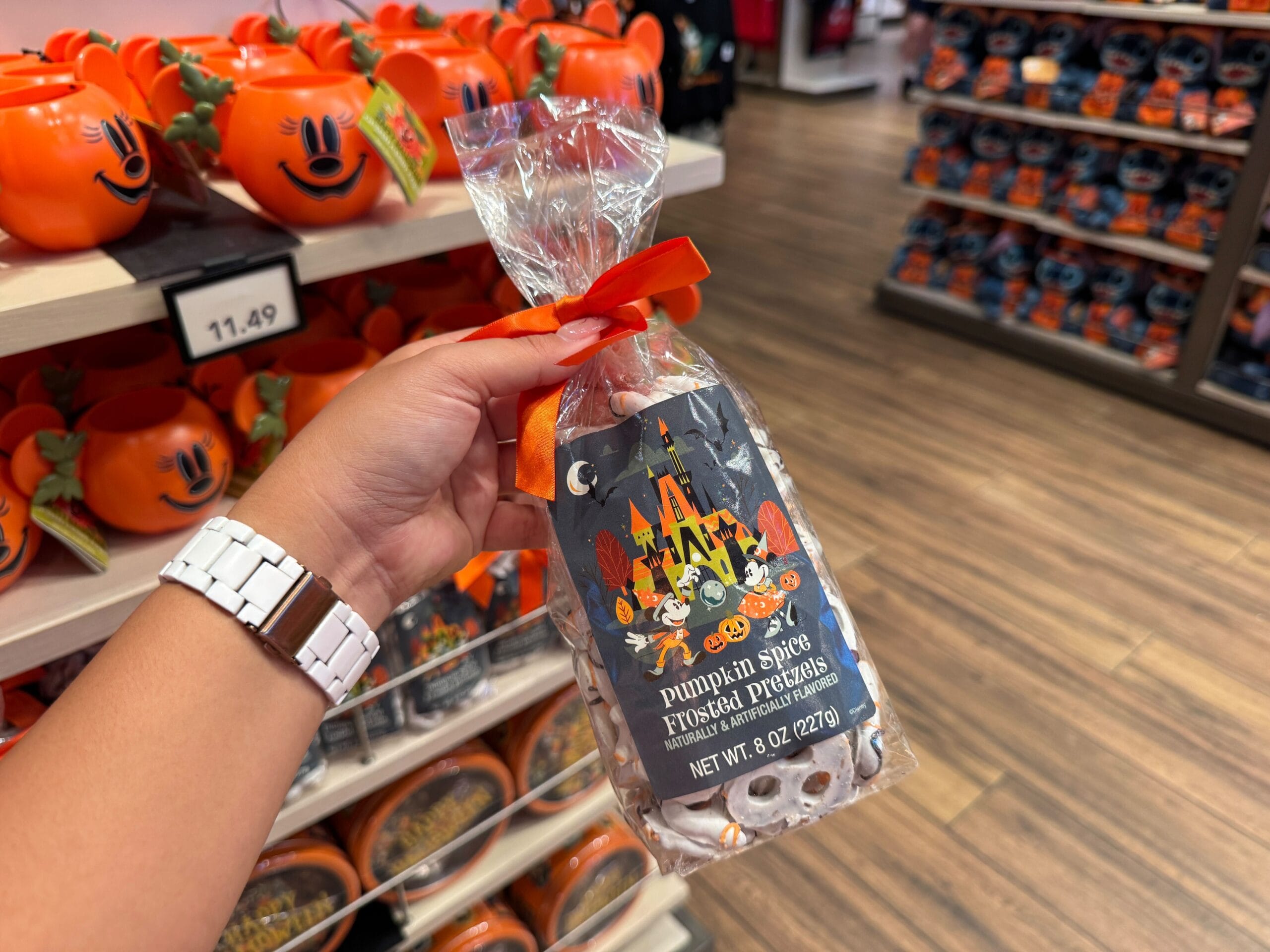 A hand holds pumpkin spice frosted pretzels in a store with 2025 Halloween merchandise and pumpkin mugs on shelves.