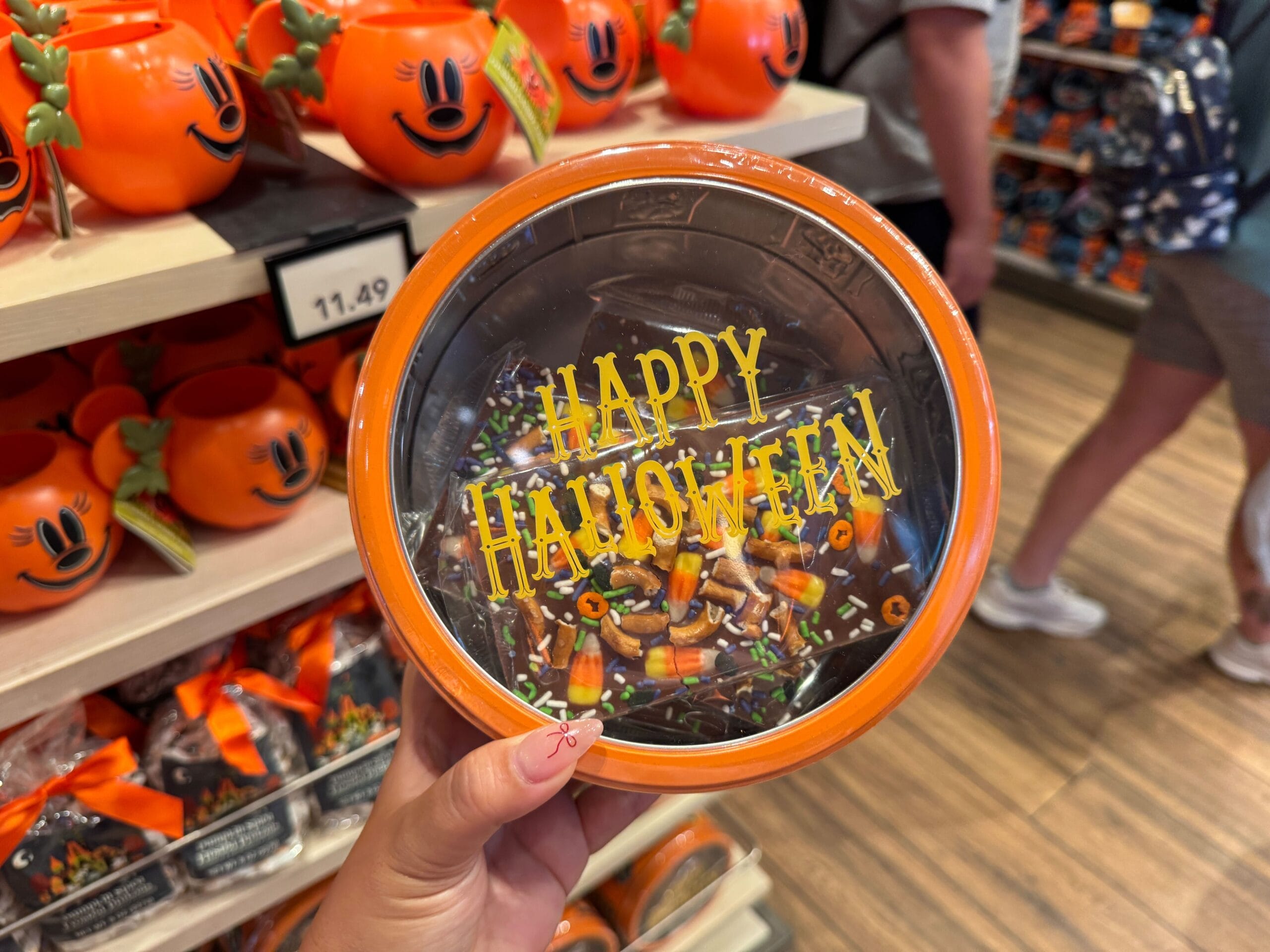 A hand holds an orange bowl with "Happy Halloween" text; 2025 Halloween merchandise fills Walt Disney World shelves.