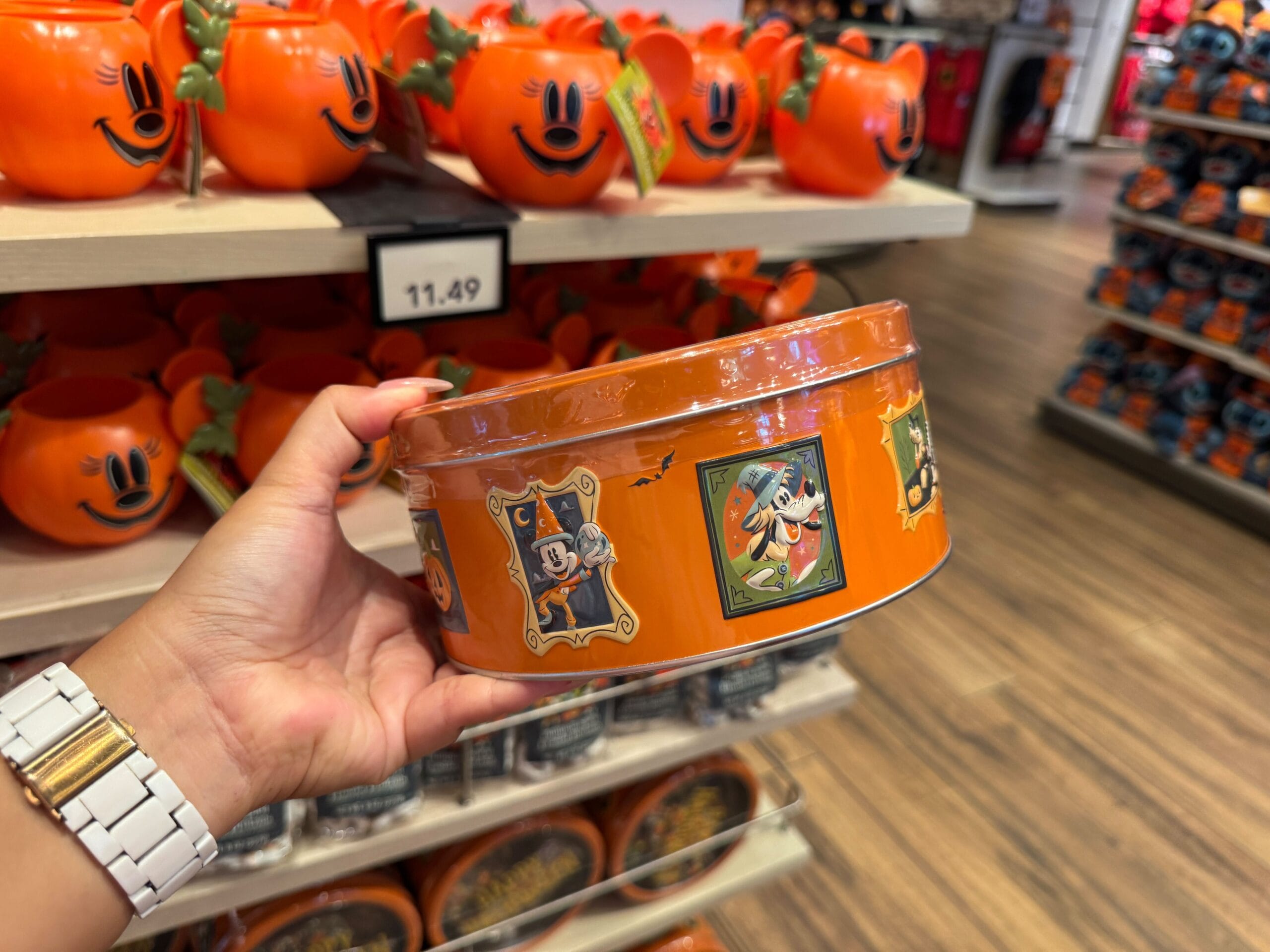 Hand holds an orange Walt Disney World Halloween tin with Mickey, surrounded by pumpkin containers on a store shelf.