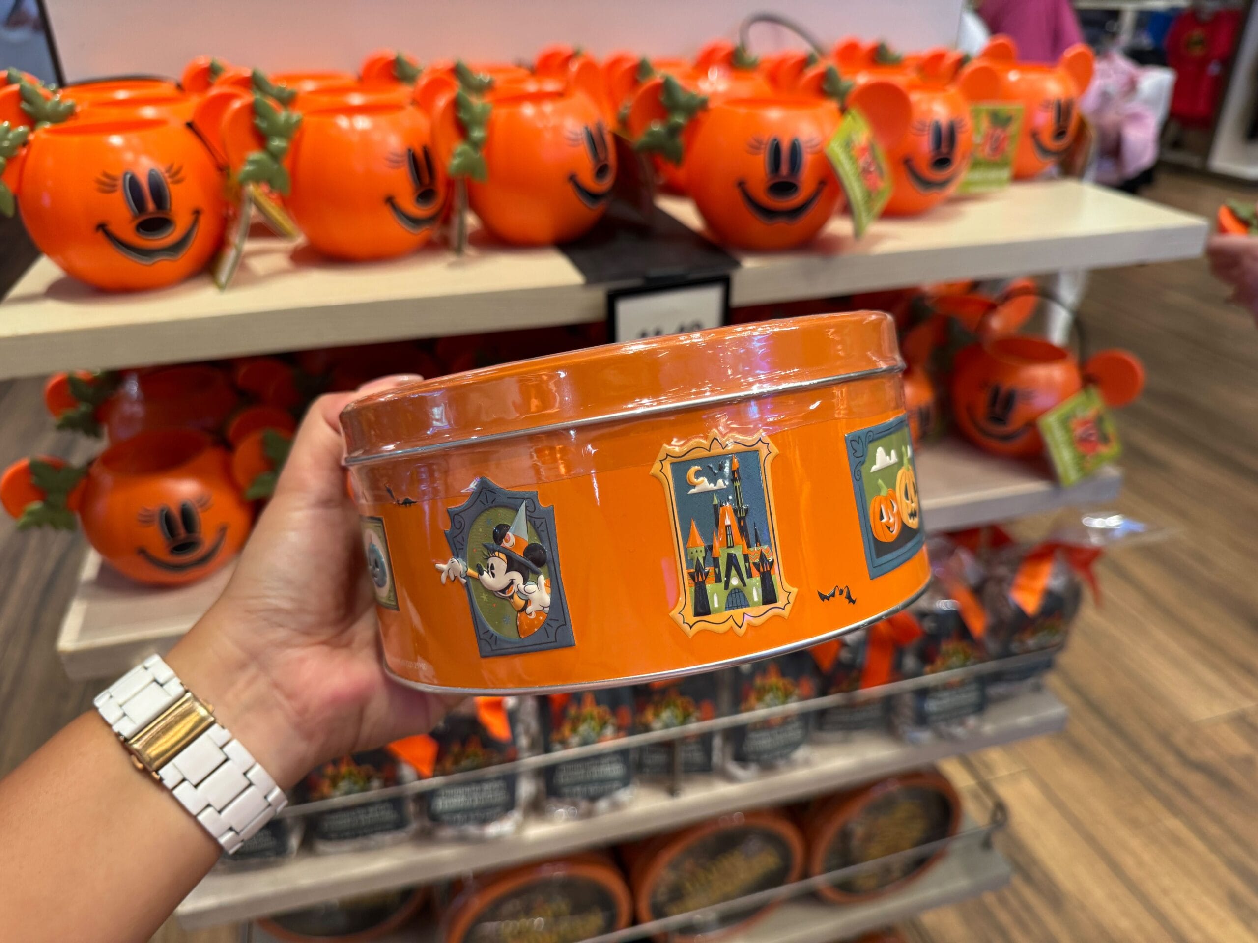 A hand holds a 2025 Halloween merchandise tin with Mickey Mouse graphics in a store with pumpkin-shaped containers.