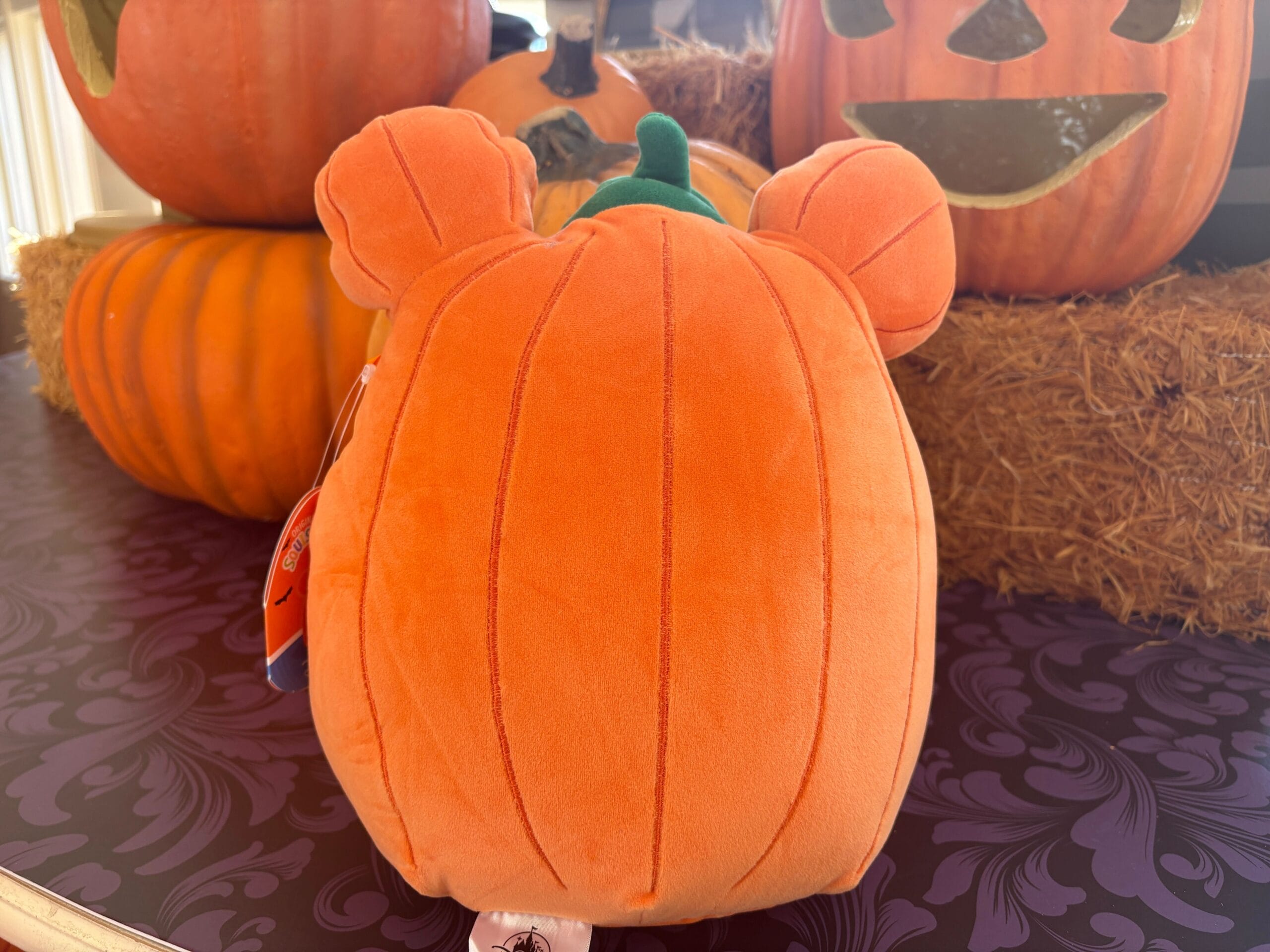 Plush pumpkin toy with bear ears, part of Walt Disney World Halloween merchandise, among stacked pumpkins and hay.