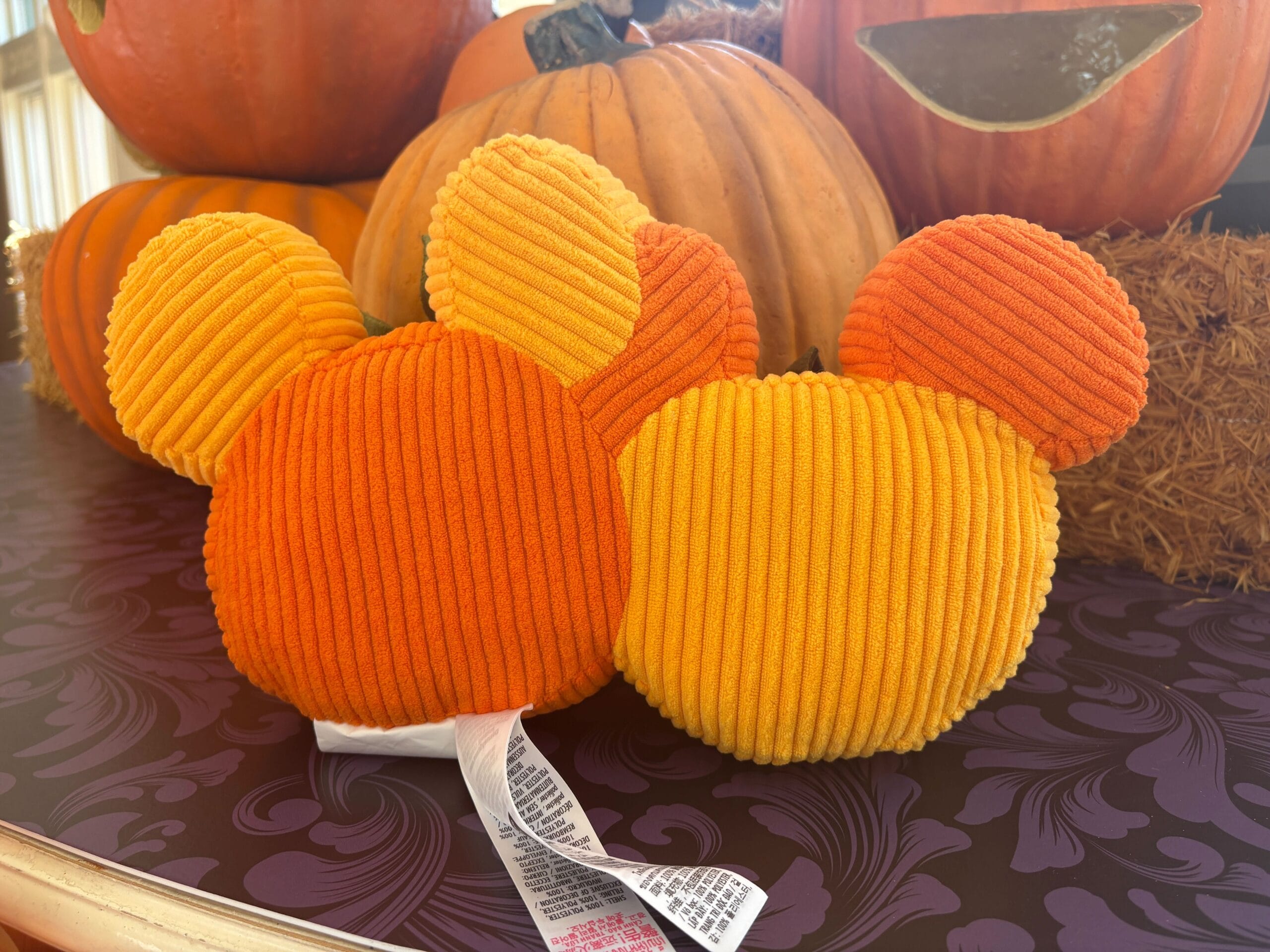 Plush pumpkin Mickey Mouse ears, part of Walt Disney World Halloween merchandise, sit among pumpkins and hay.